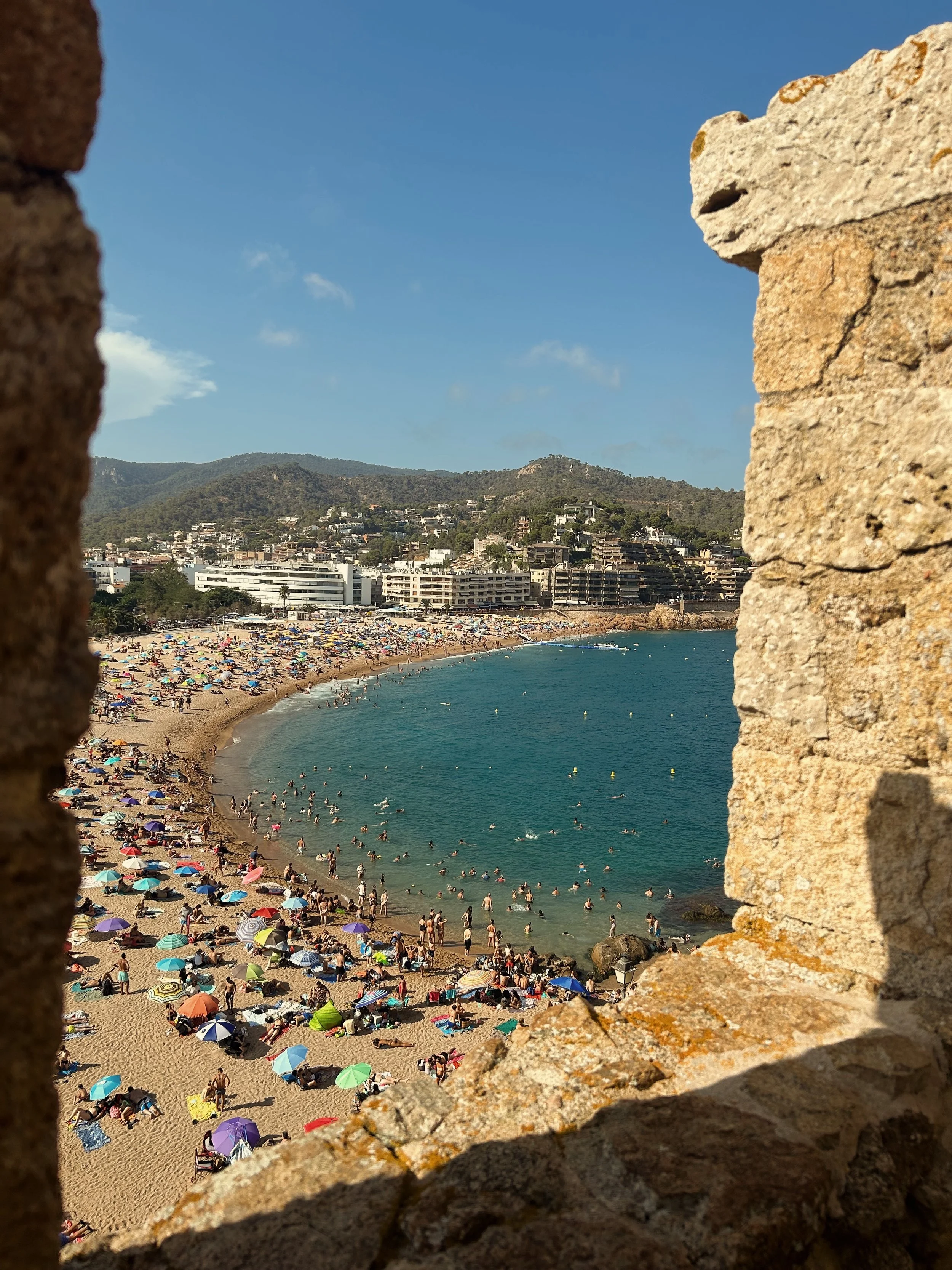Beach filled with colorful umbrellas, people swimming and sunbathing, overlooking the ocean with a cityscape and hills in the background, viewed through a stone window.
