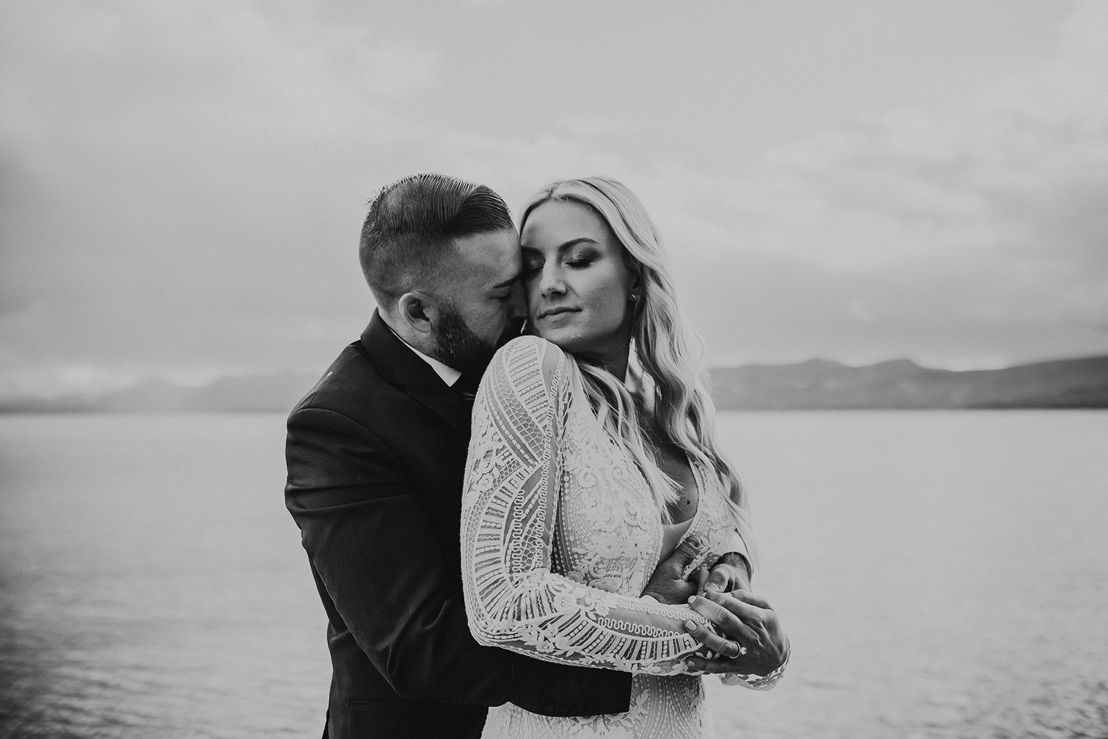 A black and white photo of a couple embracing by a body of water with mountains in the background. The woman has long wavy hair and is wearing a lace dress, while the man has a beard and is wearing a suit. They are close with their eyes closed.