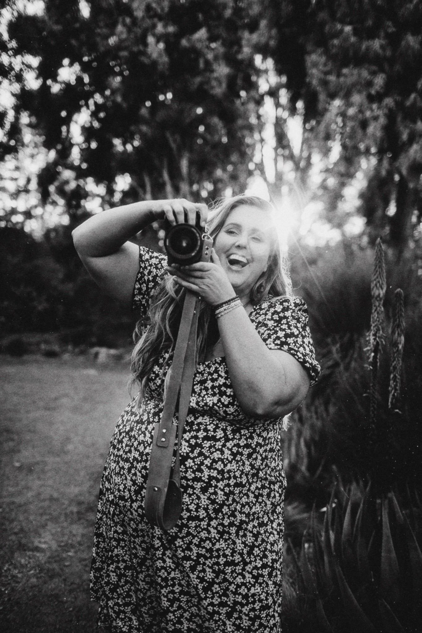 A woman smiling and holding a camera, taking a selfie outdoors at sunset with trees in the background.