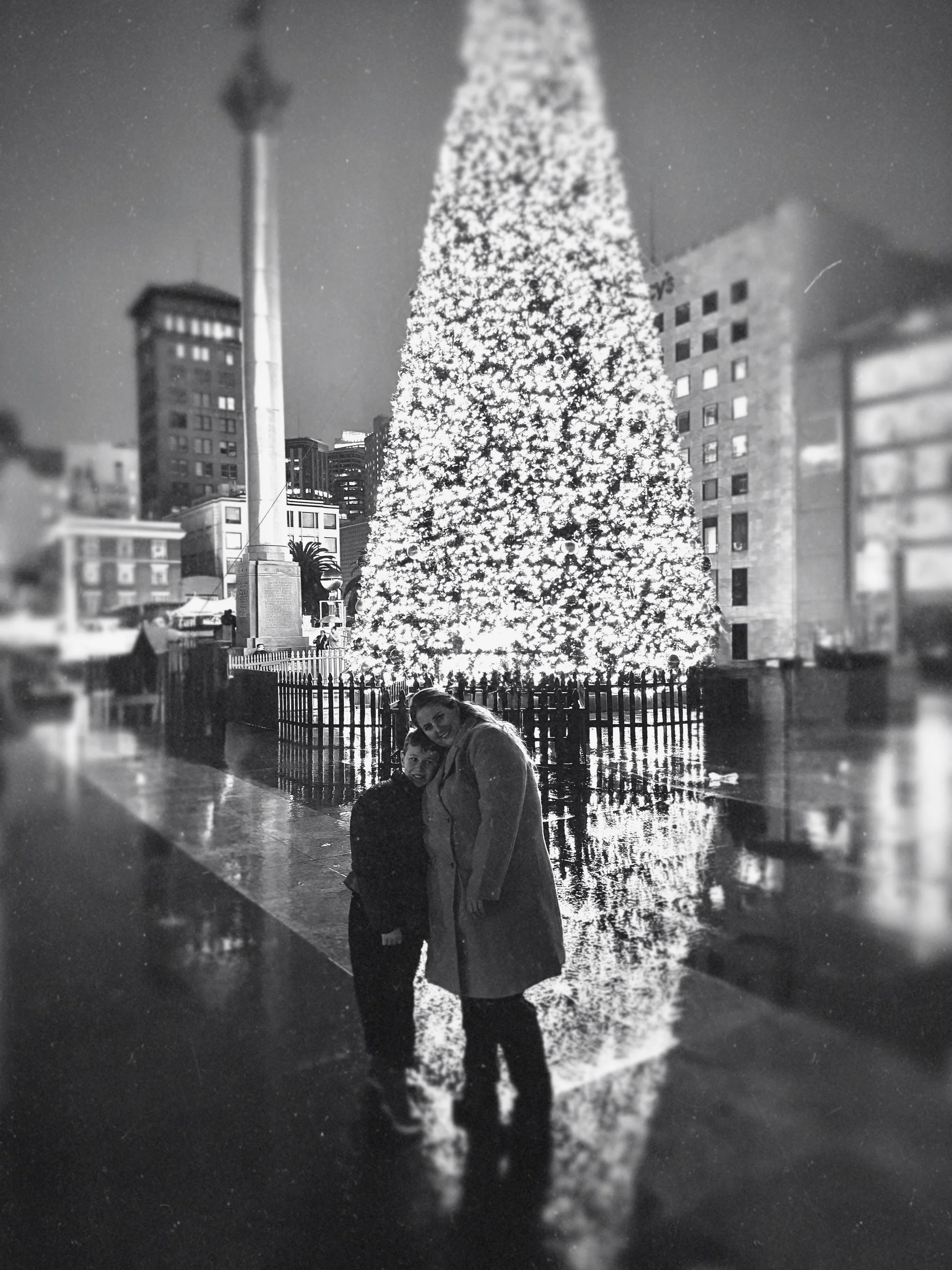 A woman and a boy standing together in front of a large, illuminated Christmas tree on a city street at night, with tall buildings and a smoking chimney in the background.