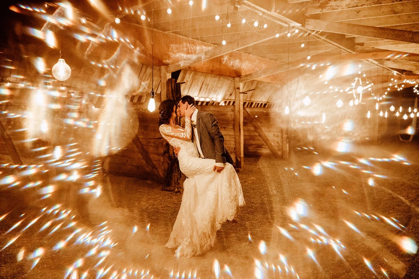 A bride and groom kissing in a warmly lit barn with string lights and hanging bulbs, surrounded by a glow and bokeh effect.