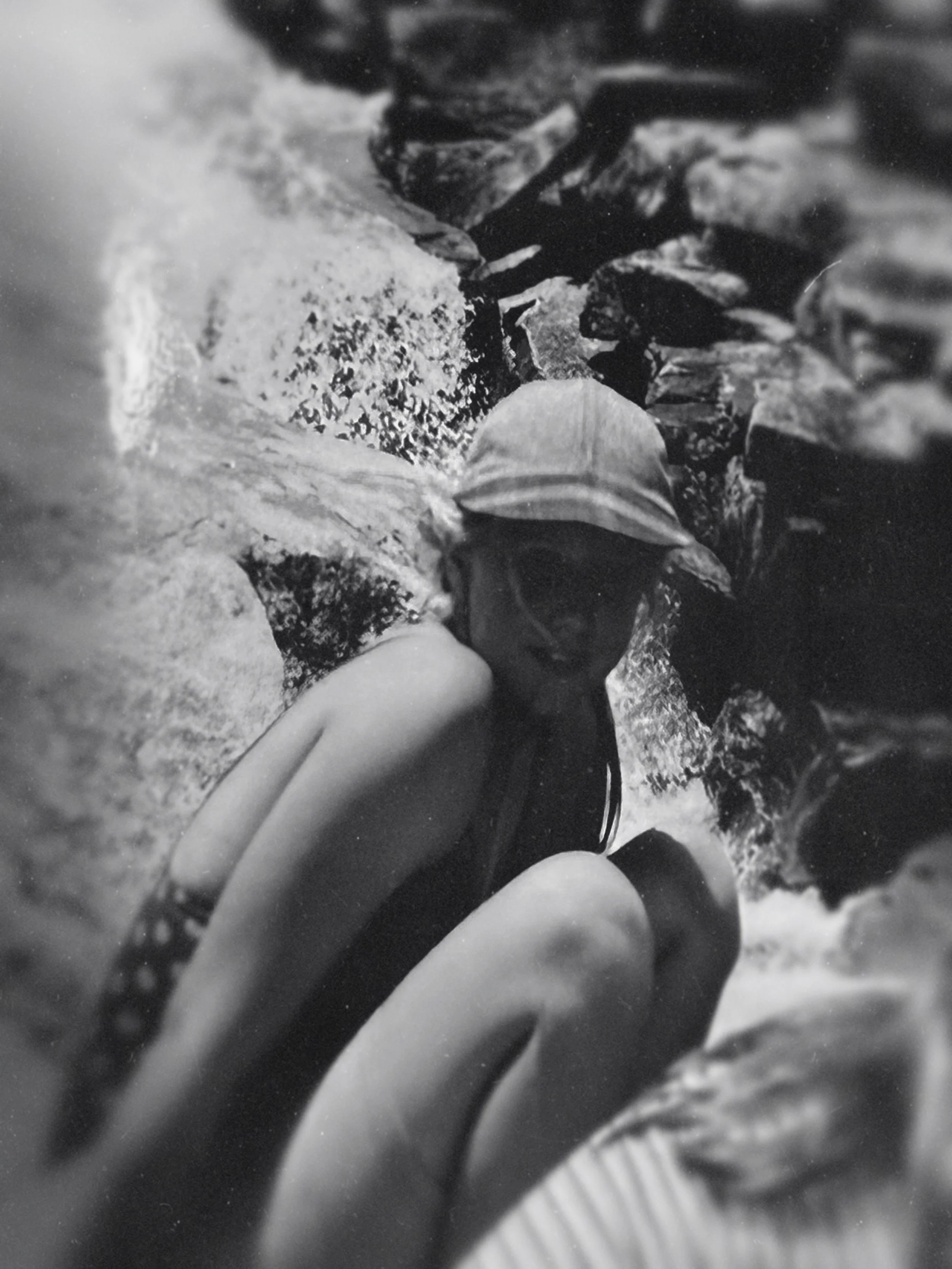 A person wearing a swimming cap and goggles, crouched between rocks and water, in a river or stream.