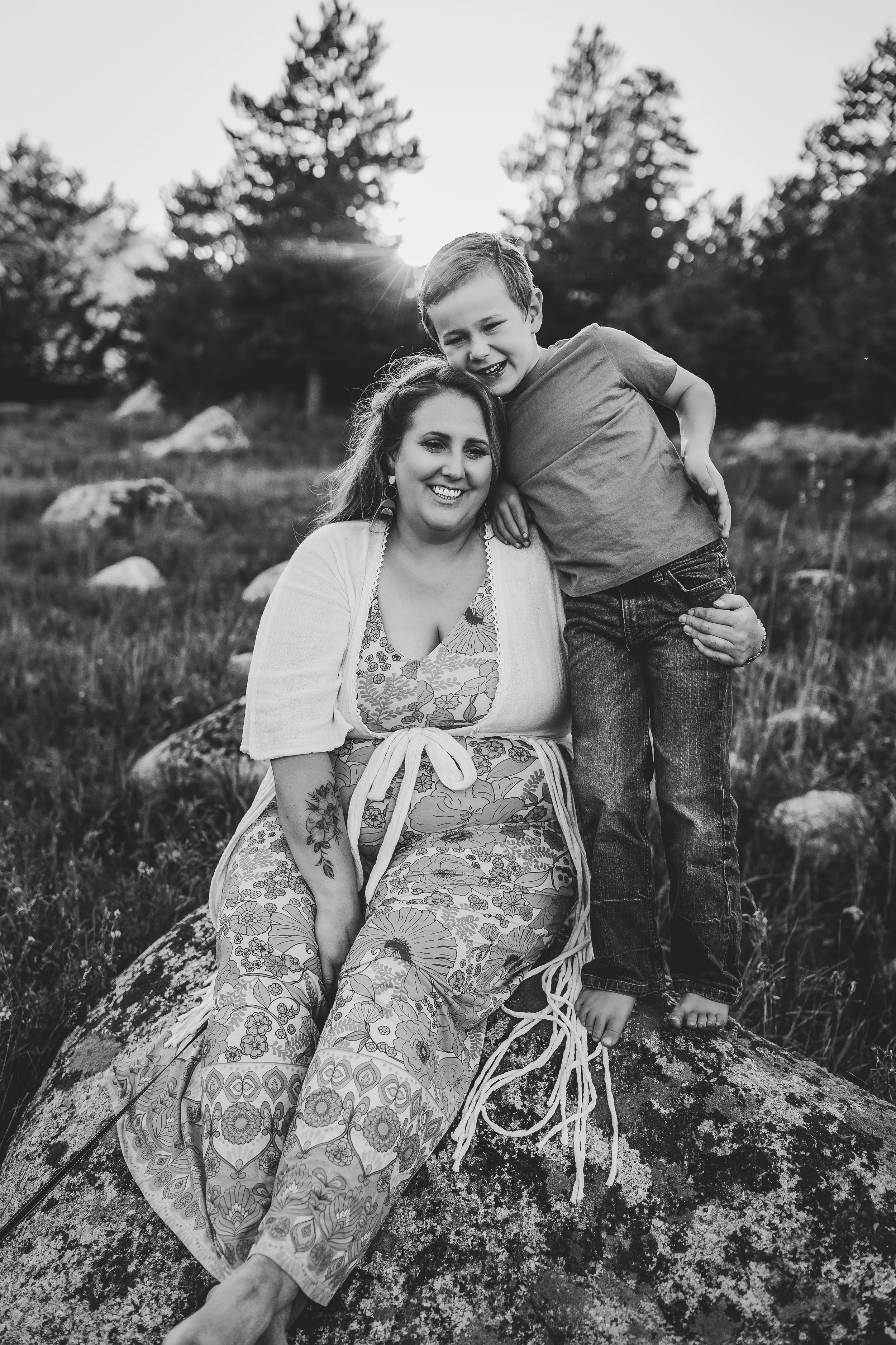 A woman and a young boy sitting and standing on a large rock outdoors, smiling, with trees and rocks in the background, in black and white.