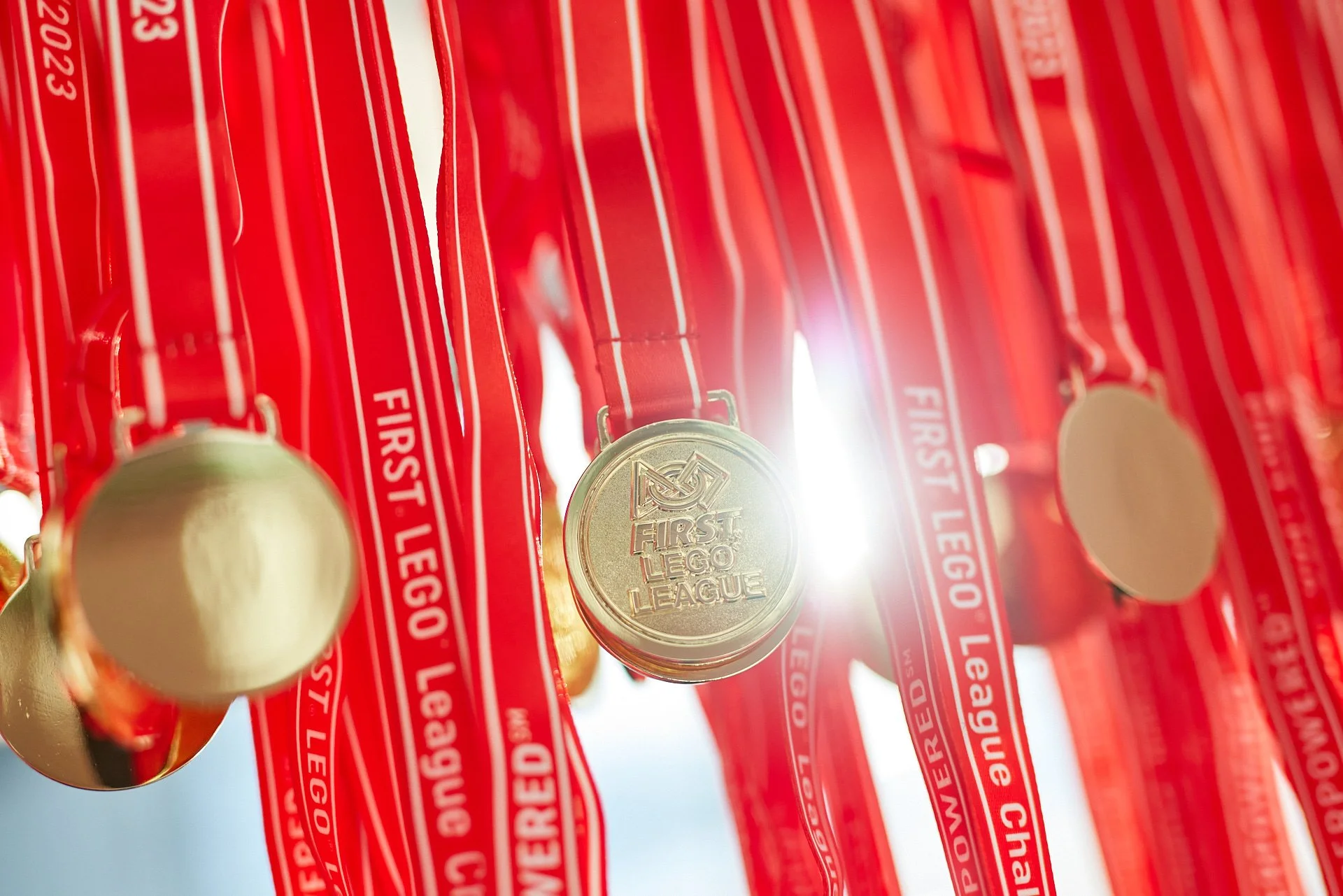 Mehrere goldene Medaillen mit dem Logo 'First Lego League' hängen an roten Bändern. Die Medaillen sind auf einem weißen Hintergrund.