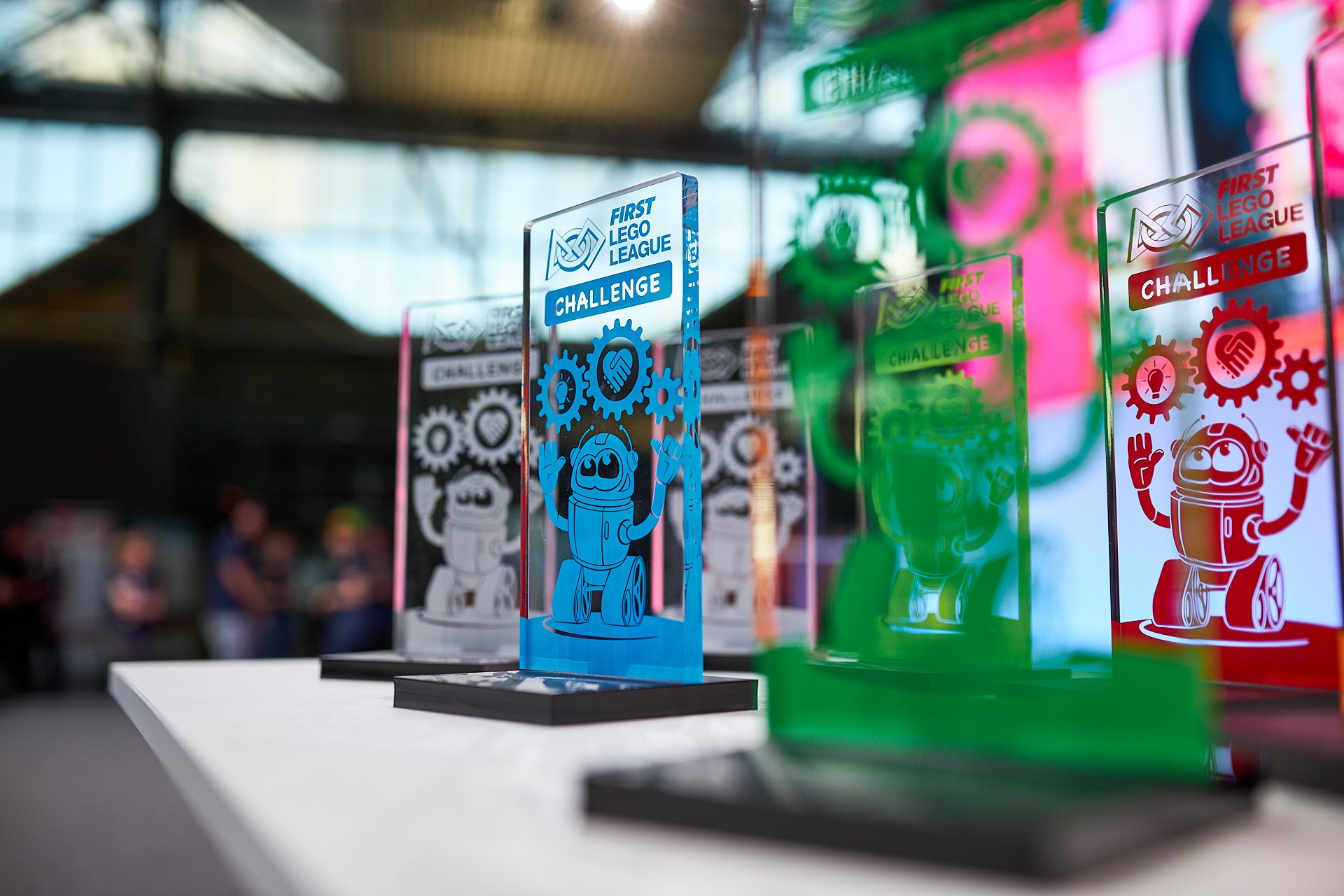 Preise für die FIRST LEGO LEAGUE Challenge auf einem Tisch, bunte Glas-Trophäen mit LEGO Robotern und Gears, im Hintergrund Besucher bei einer Veranstaltung.