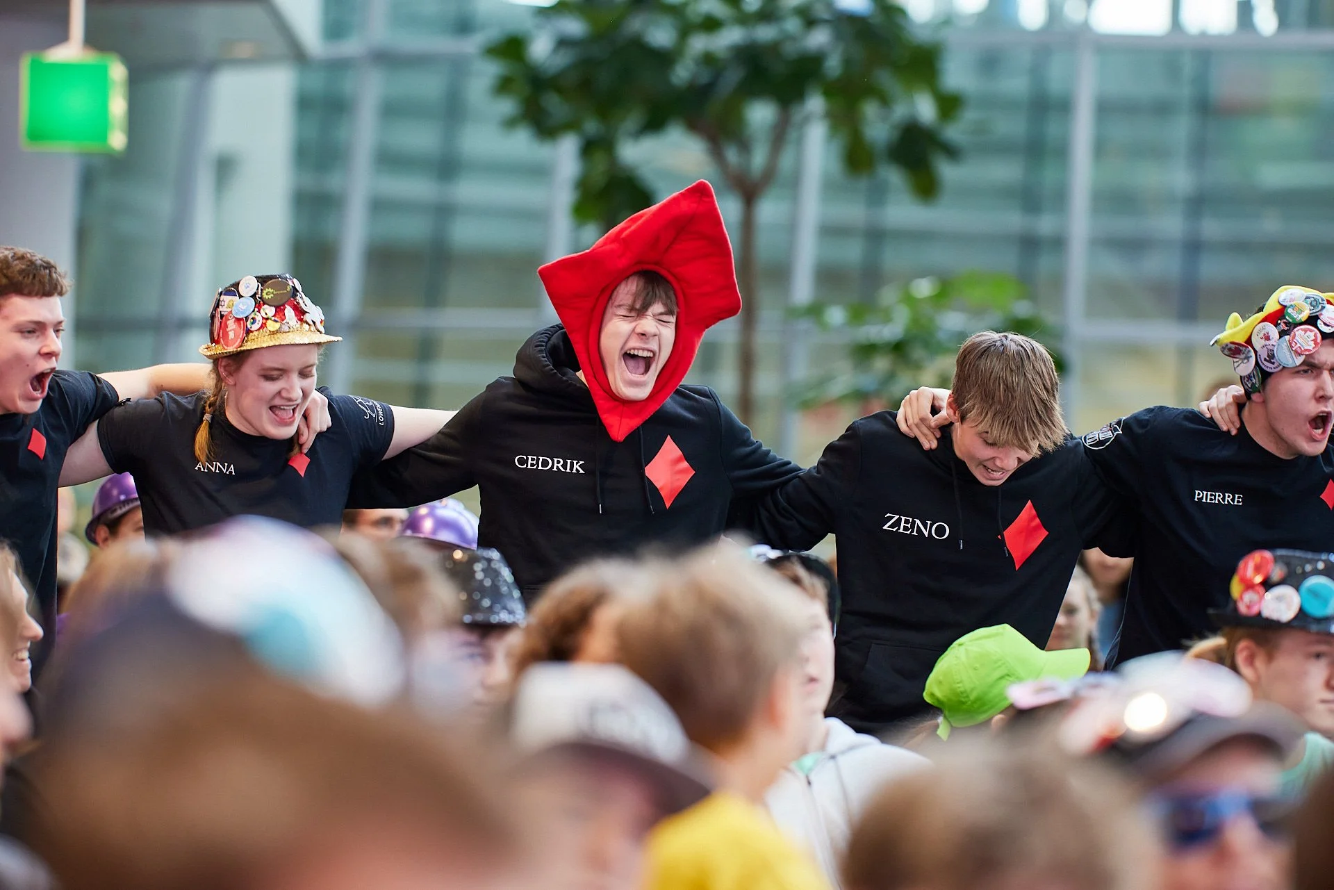 Gruppen von lachenden Jugendlichen, die bei einer Veranstaltung zusammenstehen, einige tragen bunte Hüte und Kostüme, im Hintergrund ein moderner, verglaster Gebäudeteil.