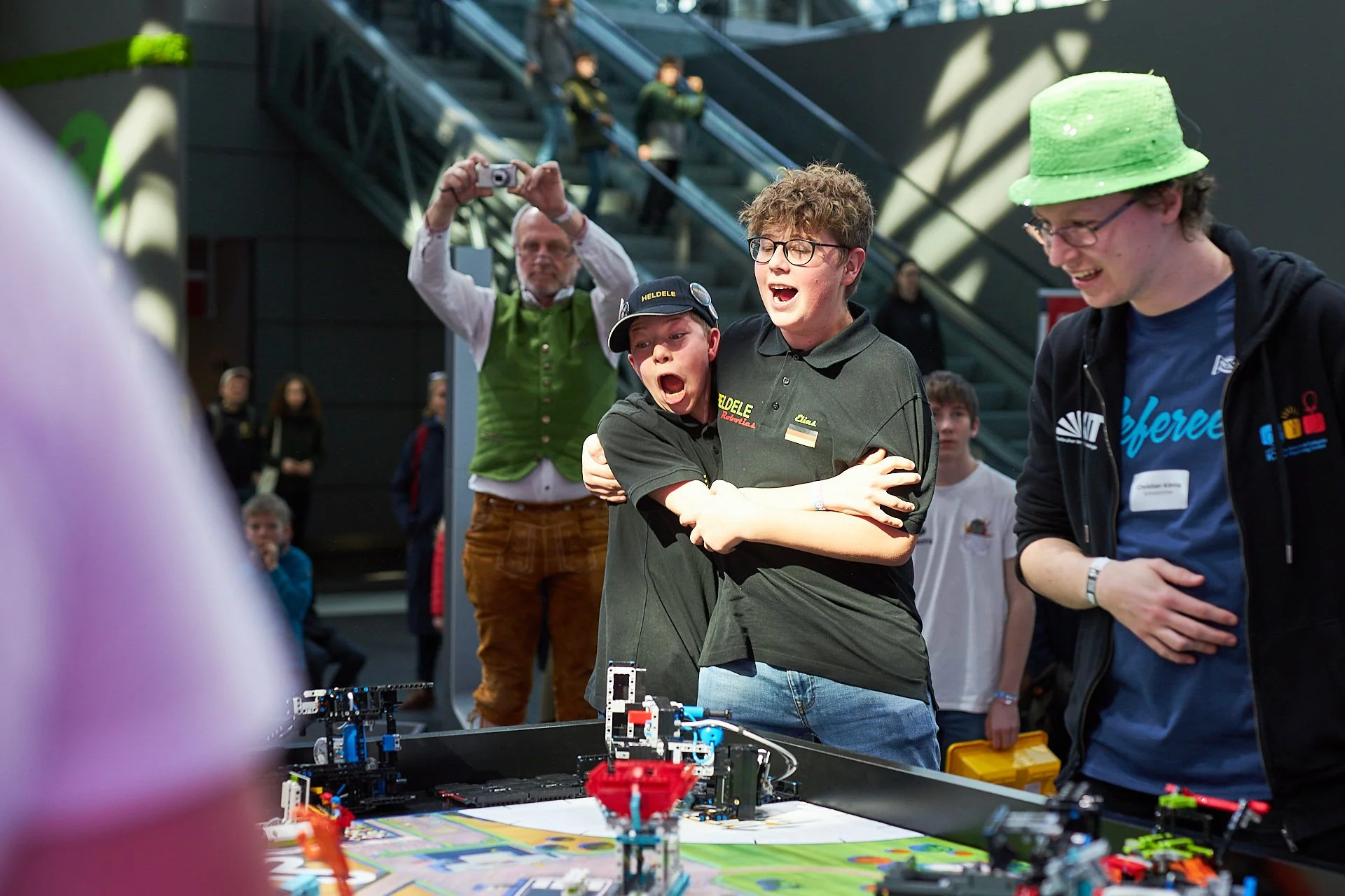 Junge bei einem Robotik-Wettbewerb schreit und umarmt einen anderen Jungen, während ein Mann in grünem Outfit und eine Person mit grünem Hut zuschauen; im Hintergrund another Personen, some taking photos.