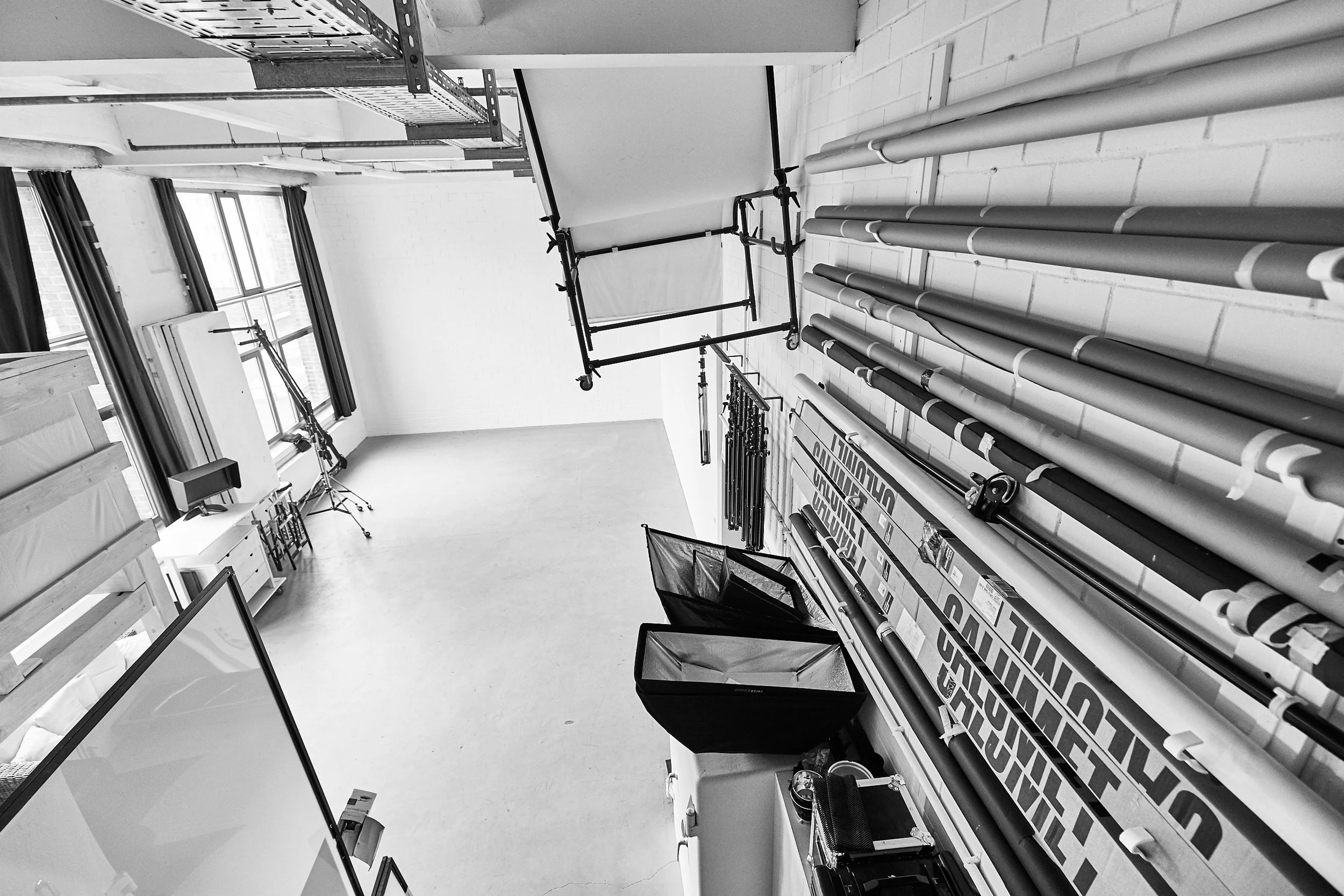 Ein Raum mit großen Fenstern, Regalen und Sportausrüstung, darunter Latten, Trockengestelle und Polstermatten, alles in Schwarz-Weiß-Fotografie.