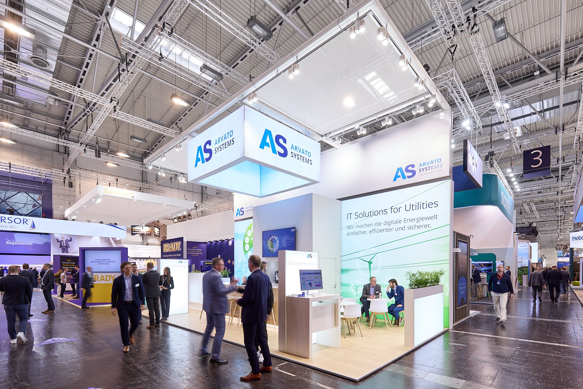 Ausstellungsstand bei einer Messe, hervorgehoben ist die Firma ARVATO SYSTEMS mit einem weißen Stand, auf dem mehrere Gesprächsteilnehmer zu sehen sind. Das Standdesign ist modern mit weißen Flächen, Bildschirmen und grünen Akzenten.
