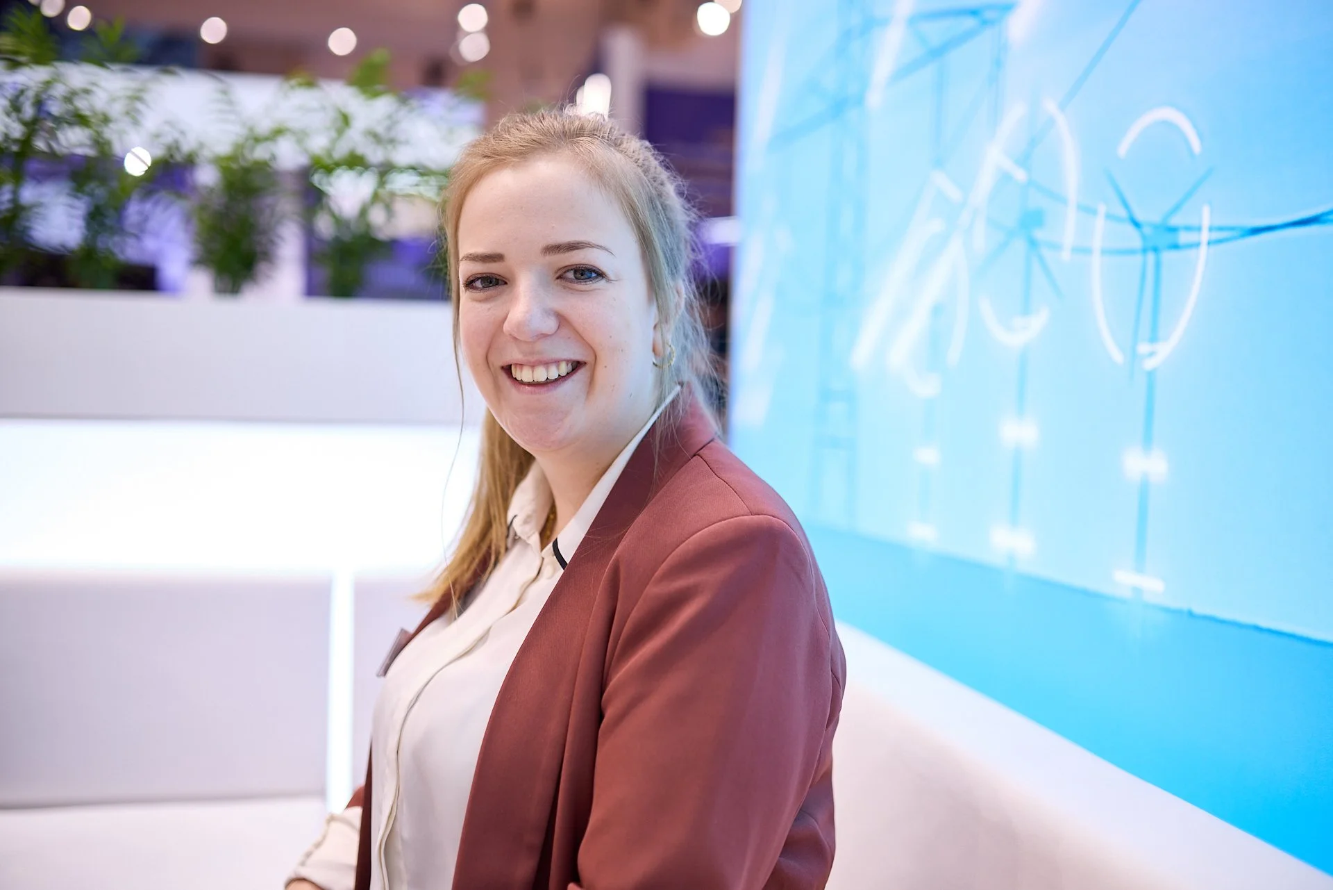 Eine lächelnde Frau in einem braunen Blazer sitzt in einem modernen Büro oder Konferenzraum, im Hintergrund eine blaue Tafel mit weißen Skizzen.