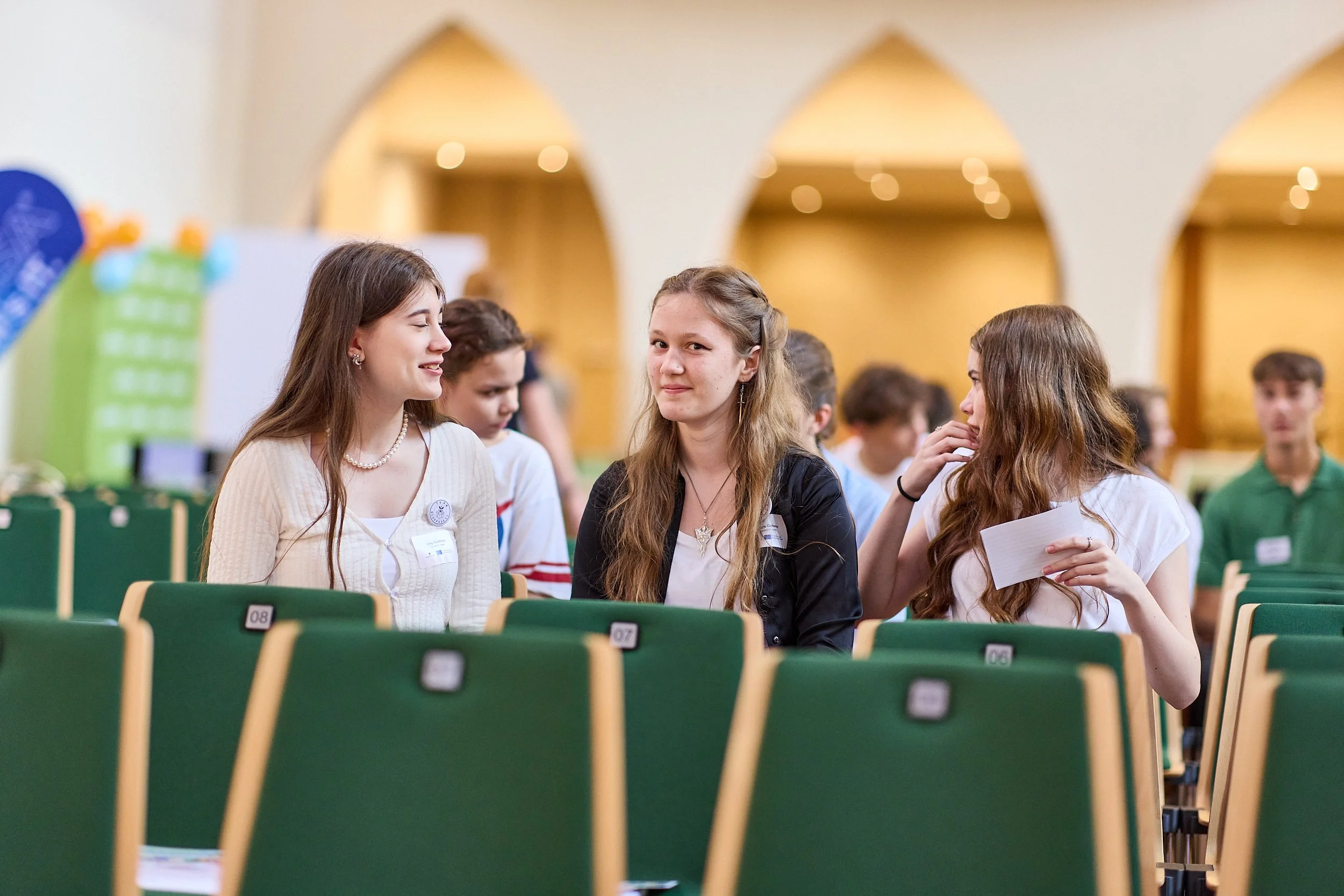 Drei junge Frauen sitzen in einem großen Raum mit grünen Stühlen, sie scheinen ein Gespräch zu führen. Im Hintergrund sind andere Jugendliche und die Räumlichkeiten mit Holzdekoren sichtbar.