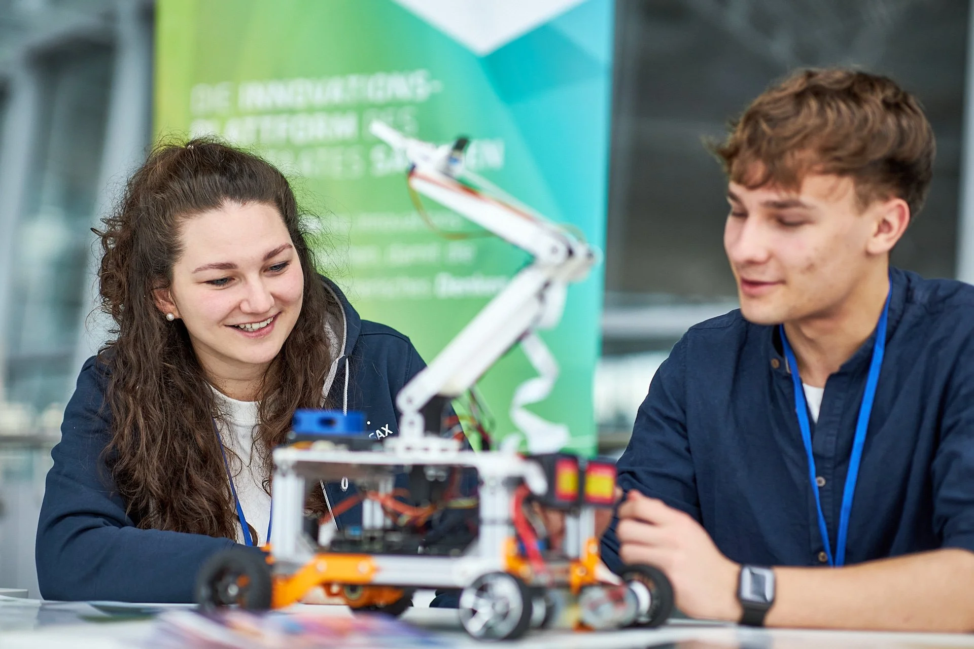Zwei junge Menschen, eine Frau und ein Mann, schauen lächelnd auf einen selbstgebauten Roboter mit Greifarm, der auf einem Tisch steht.