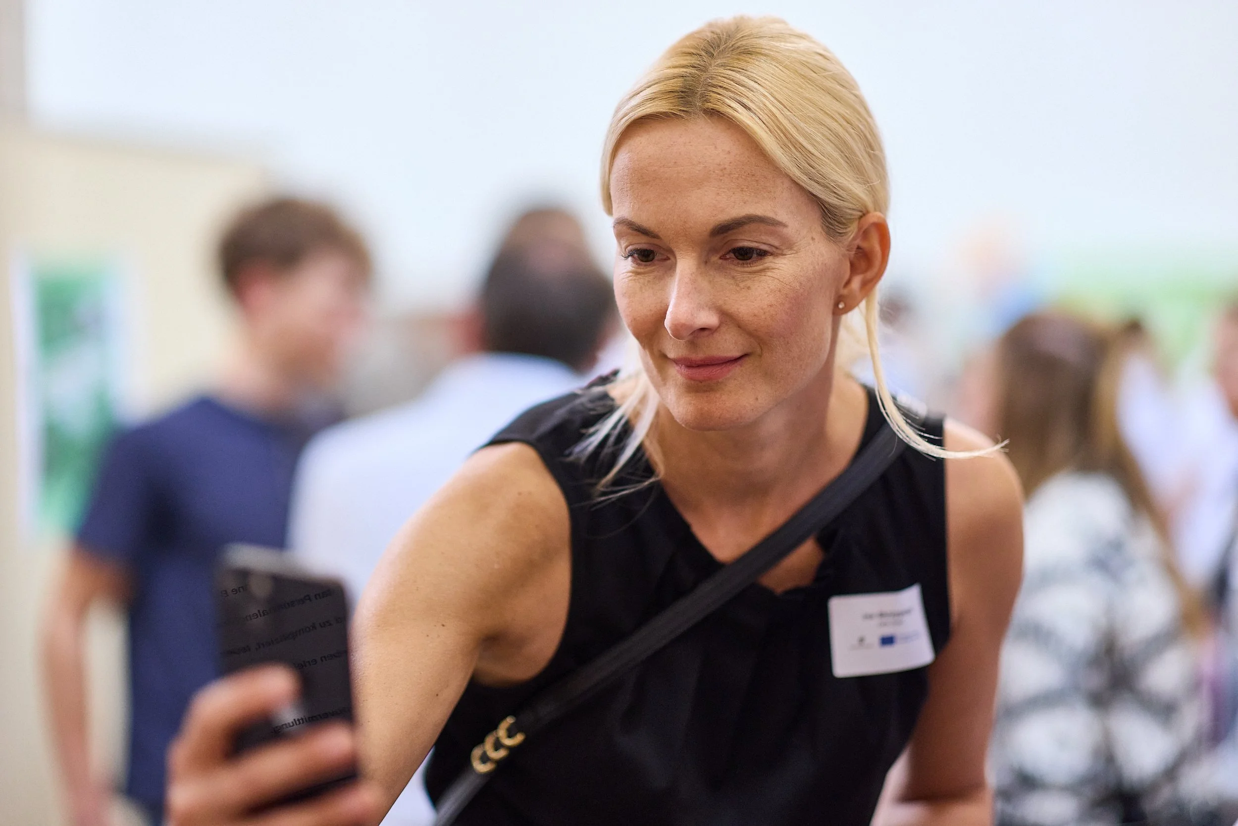 Eine blonde Frau macht Selfie auf einer Veranstaltung mit vielen Menschen im Hintergrund.