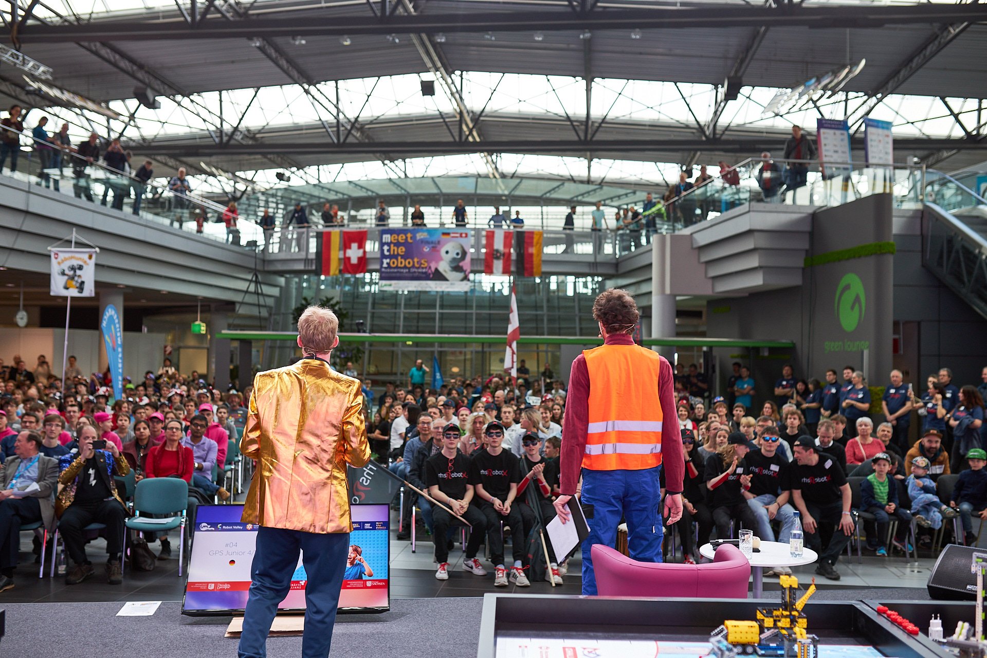 Zwei Moderator vor einem Publikum bei einer Robotik-Veranstaltung in einem großen, modernen Gebäude, mit Flaggen und einem Banner im Hintergrund.