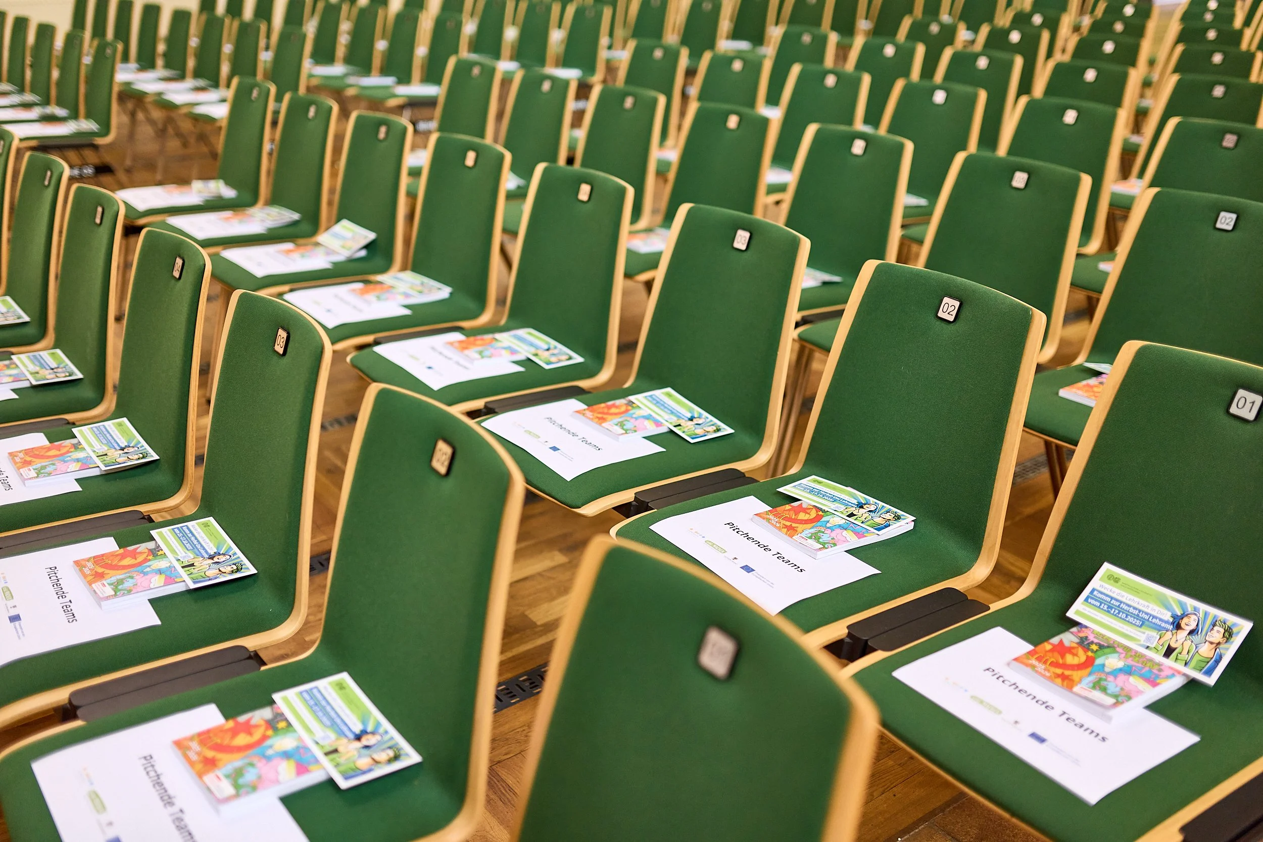 Leere grüne Sitzplätze mit Unterlagen und Flyern für eine Veranstaltung, in einem großen Saal.