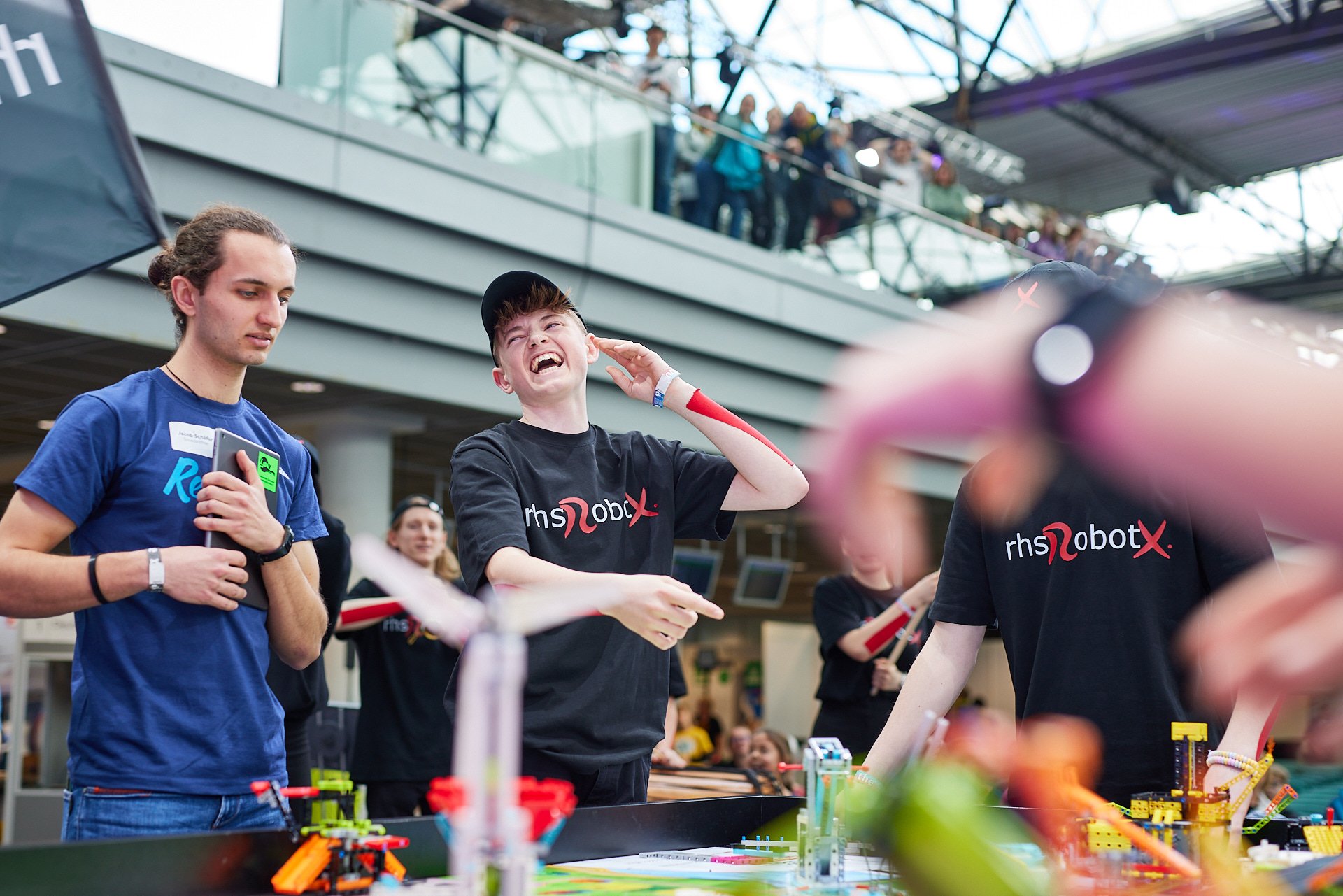 Jugendliche bei einem Robotics-Wettbewerb in einer großen Halle, das zentrale Mädchen lacht und hält Hände an die Ohren, während die anderen um einen Roboter und eine Spielplatte herumstehen.