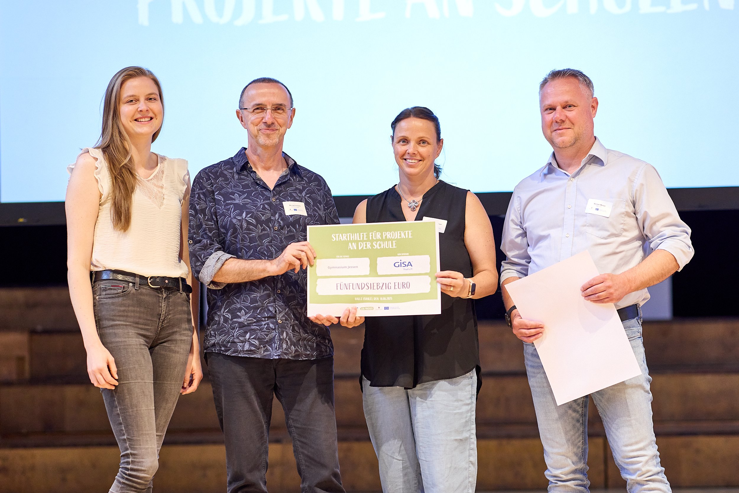 Vier Personen bei einer Preisverleihung halten eine große Quittung mit 5.059 Euro. Die Personen stehen auf einer Bühne, im Hintergrund ein Bildschirm mit Text. Die Stimmung ist freudig und formell.