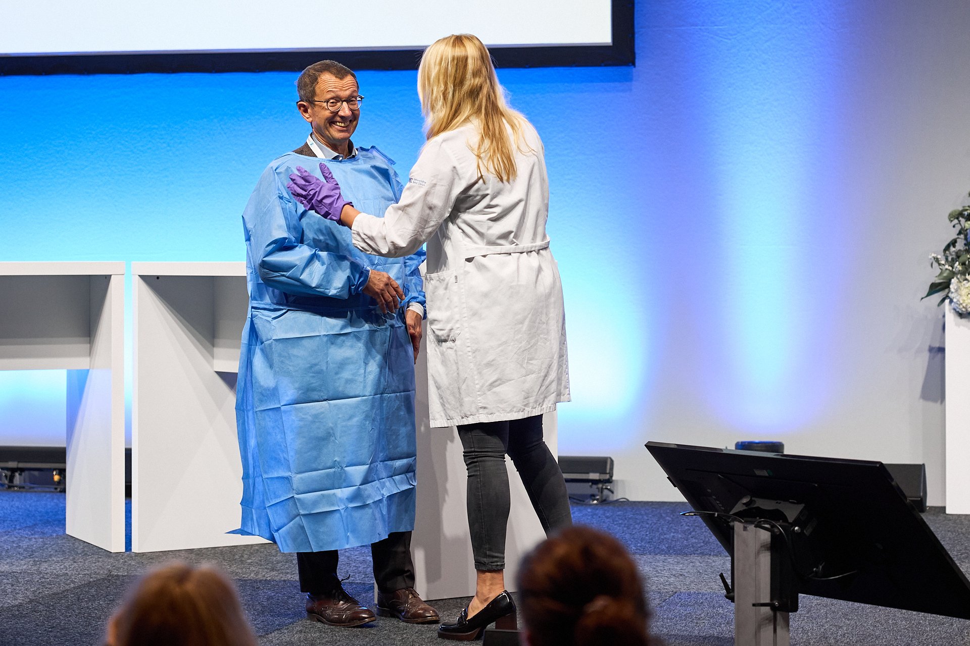 Zwei Personen, ein Mann in blauer Schutzkleidung und eine Frau in einem weißen Arztmantel, stehen auf einer Bühne bei einer Zeremonie. Sie begrüßen sich mit einem Handschlag, beide lächeln.
