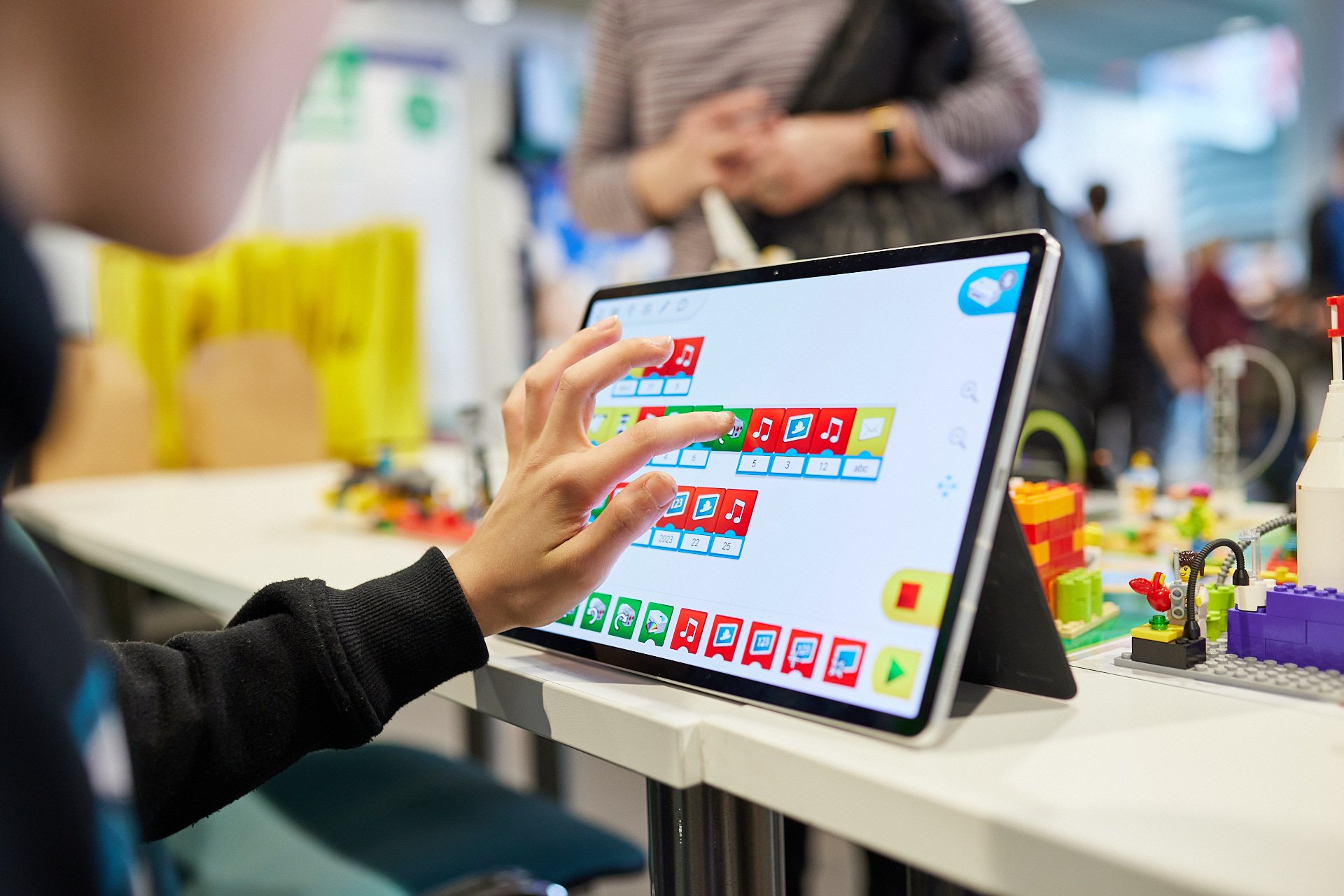 Person interagiert mit einem Tablet, das ein Programm mit bunten Symbolen und Blöcken anzeigt, wahrscheinlich beim Spielen oder Lernen mit Bausteinen, im Hintergrund sind weitere Menschen und LEGO-Teile sichtbar.