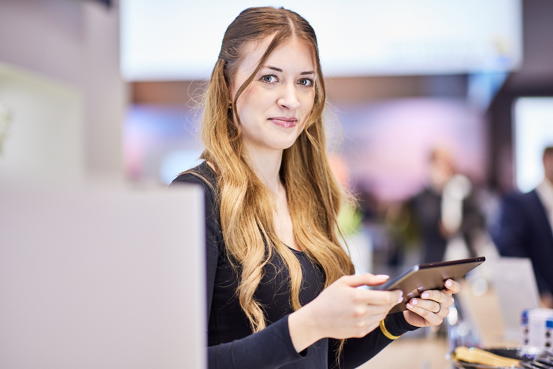 Eine junge Frau mit langem, lockigem, rotem Haar hält ein Smartphone in den Händen und schaut in die Kamera, während sie in einem modernen Büro arbeitet.