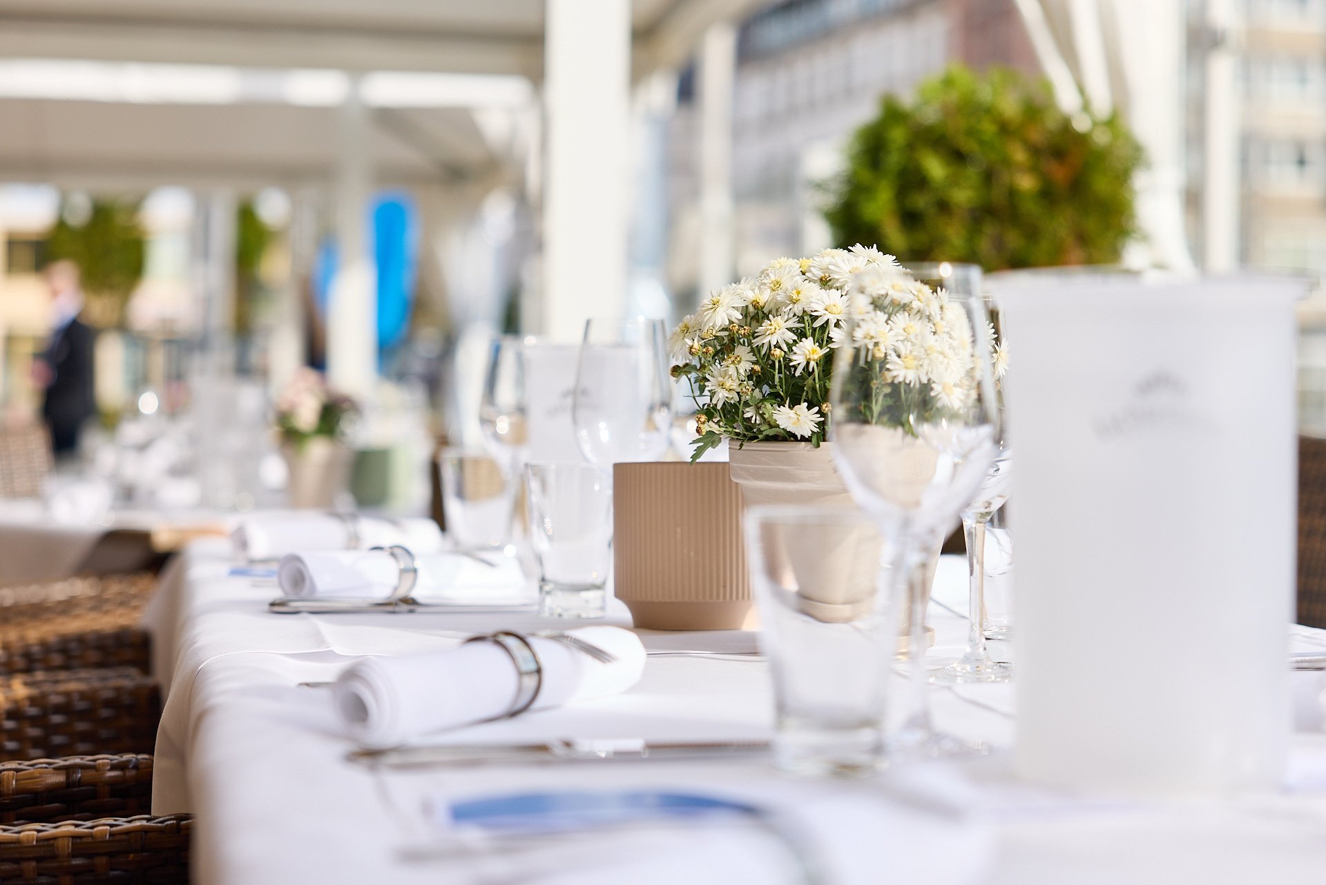 Ein festlich gedeckter Tisch mit weißen Servietten, Wein- und Wassergläsern, Blumendekoration mit weißen Blumen in Vasen, auf einem weißen Tischtuch, in einem Restaurant oder Festsaal.