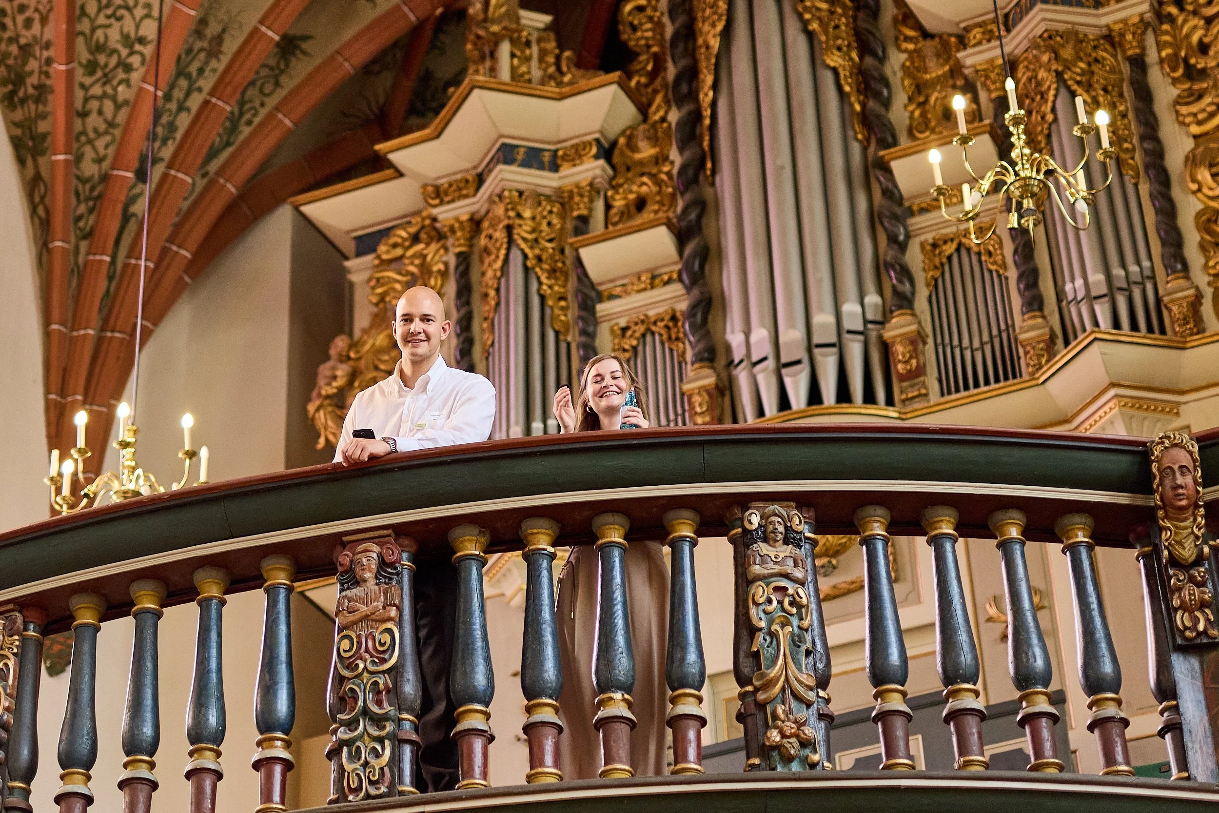 Zwei lachende Menschen, ein Mann und eine Frau, stehen auf einer Empore in einer Kirche mit barockem Orgel im Hintergrund.