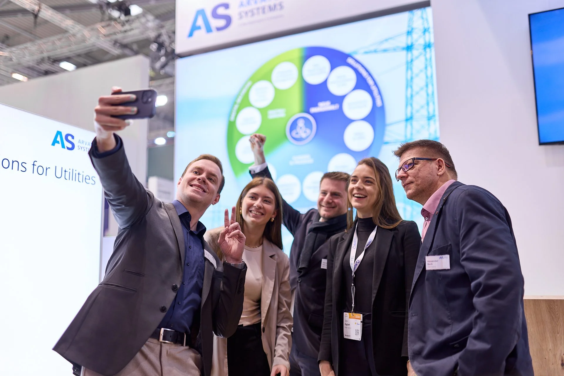 Fünf Menschen posieren für ein Gruppenfoto bei einer Messe, während sie einen Handy-Foto machen, im Hintergrund ein Bildschirm mit einem Diagramm und der Text 'AS' und 'Solutions for Utilities'.