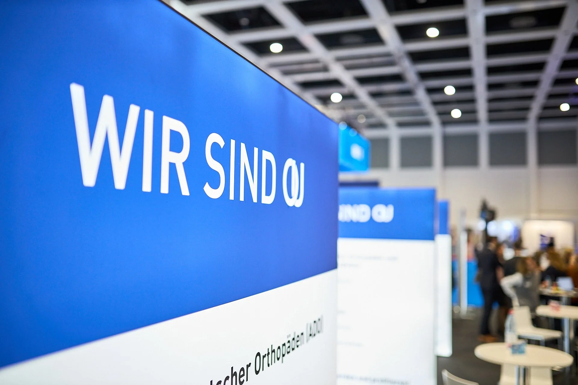 Blaue Messewand mit weißen Buchstaben, die 'WIR SIND' sagen, im Hintergrund sind weitere Messewände und Menschen, die an Ständen arbeiten und sitzen.