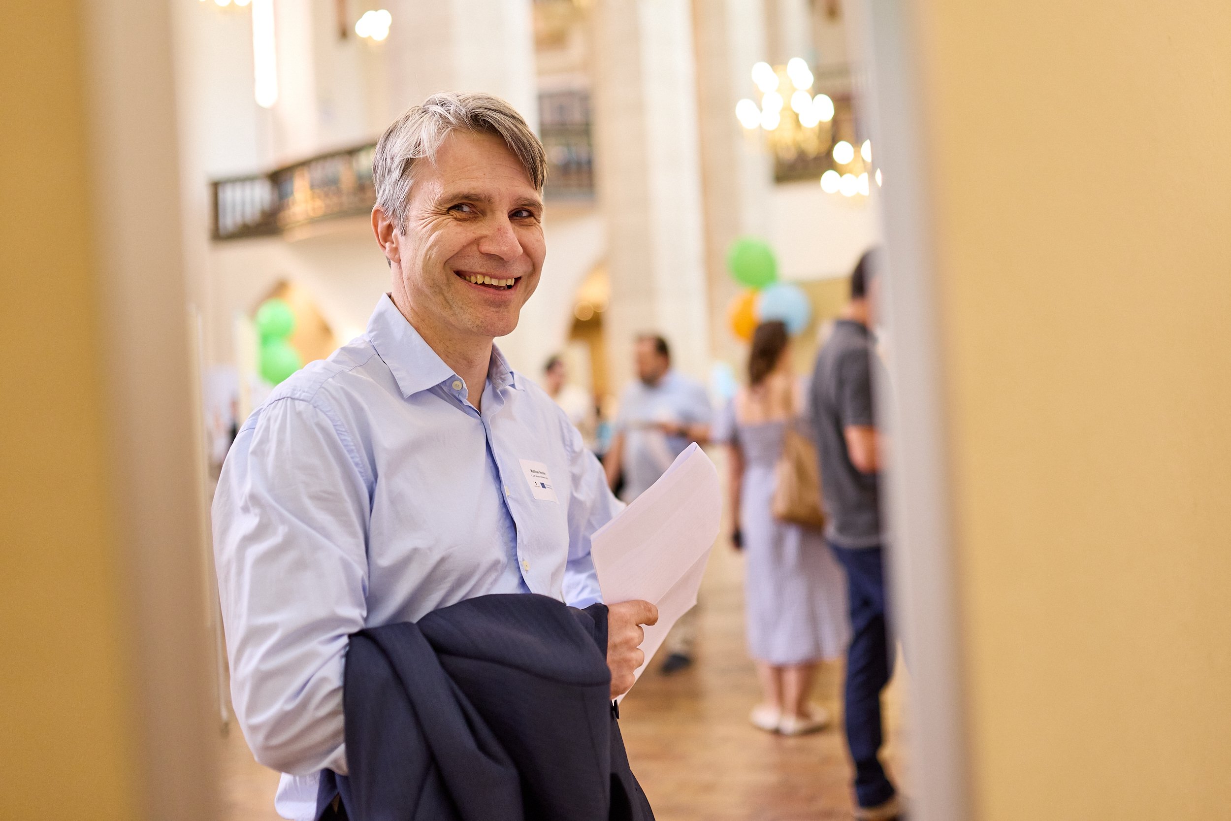 Lächelnder Mann in Hemd hält Unterlagen, bei Veranstaltung im eleganten Saal mit bekannten Dekorationen