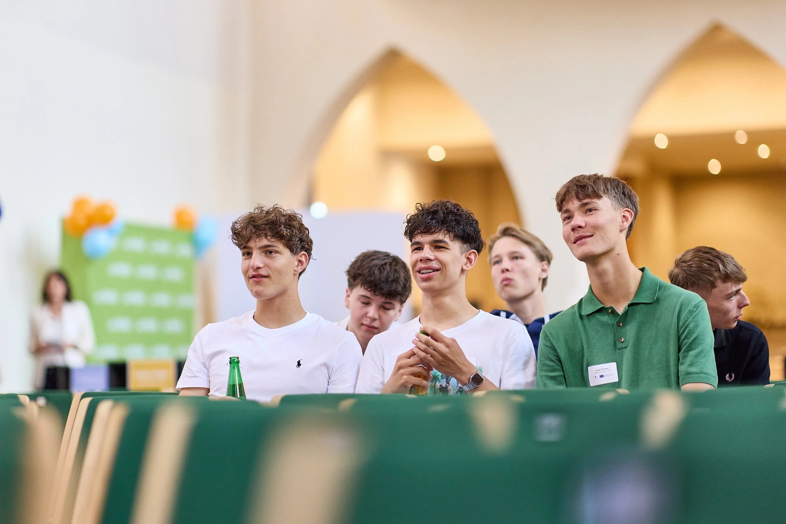 Gruppe junger Männer sitzt in einem Saal, einige lachen, andere schauen aufmerksam, bei einer Veranstaltung.
