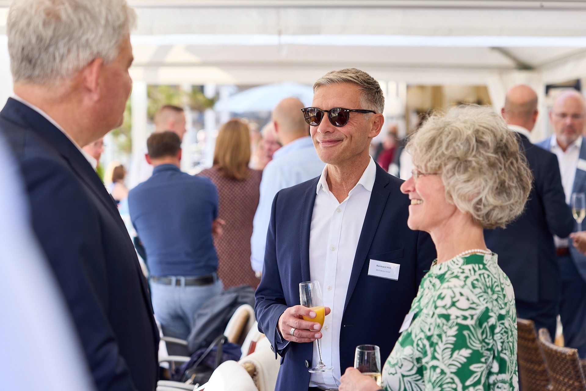 Menschen bei einer gesellschaftlichen Veranstaltung, drei Personen im Mittelpunkt, einem Mann mit dunkler Sonnenbrille, einer Frau mit Locken und einer weiteren Person im Gespräch, im Hintergrund weitere Gäste, Getränke und ein Zelt.