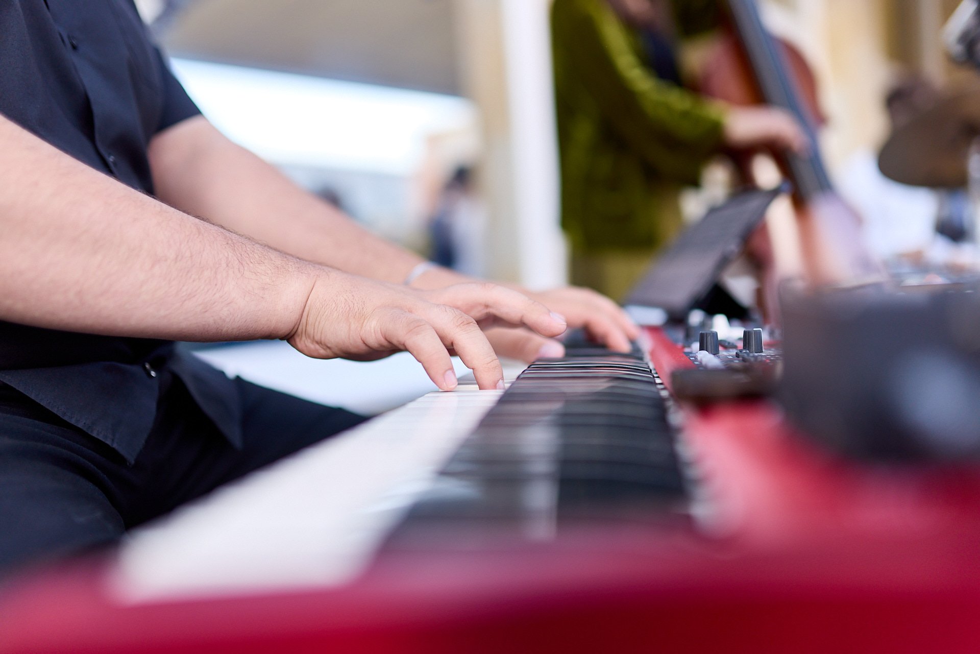 Nahaufnahme von einer Person, die auf einem elektronischen Keyboard spielt, mit weiteren Menschen im Hintergrund bei einem Konzert oder Musikveranstaltung.