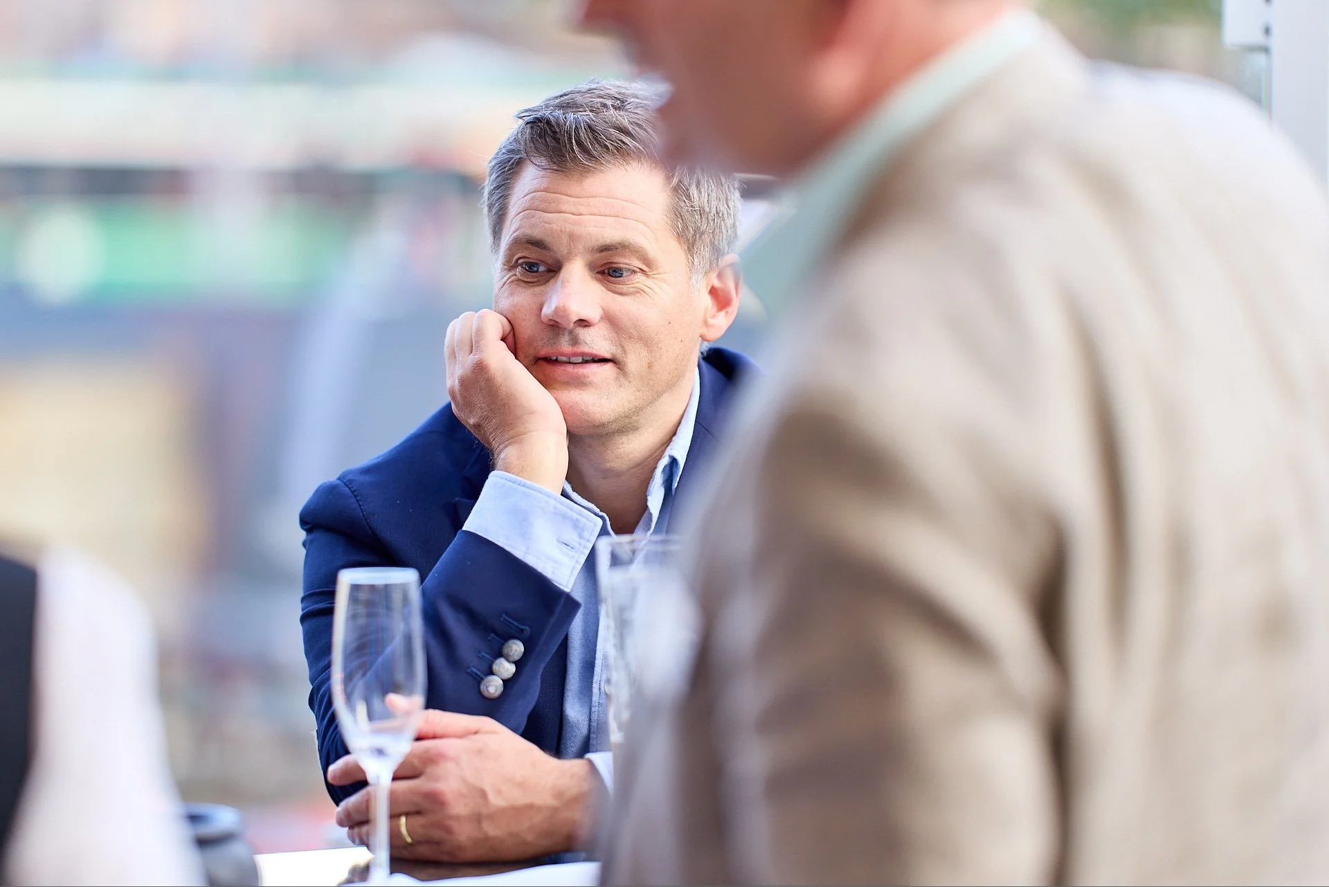 Zwei Männer im Gespräch bei einem Essen in einem Restaurant, einer sitzt mit einem Trinkglas vor sich, während der andere den Rücken zur Kamera hat.