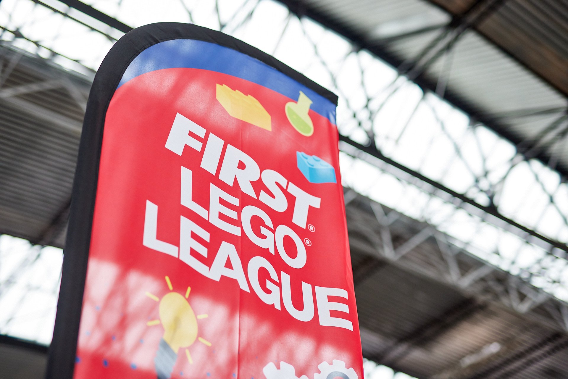 Ein rotes Banner mit dem Text "First LEGO League" in weißen Buchstaben, auf dem unterschiedliche Symbole des Lego Duplo Systems abgebildet sind, darunter eine Sonne, eine Pyramide, eine Lupe, eine Wasserblase und eine Schraube, vor einer Industriehal