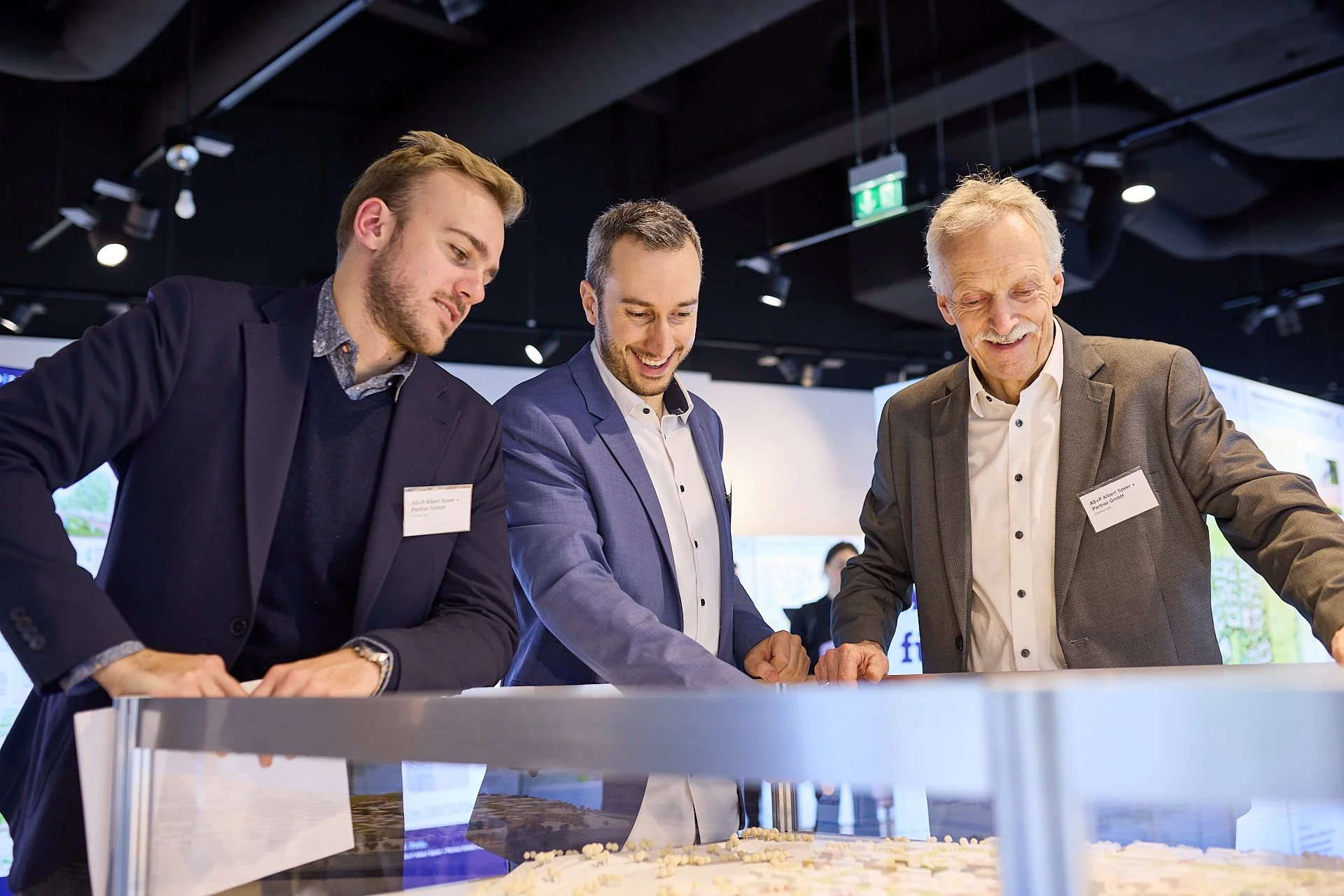 Drei Männer in Businesskleidung betrachten eine Modelllandschaft auf einem Tisch in einem Konferenzraum.