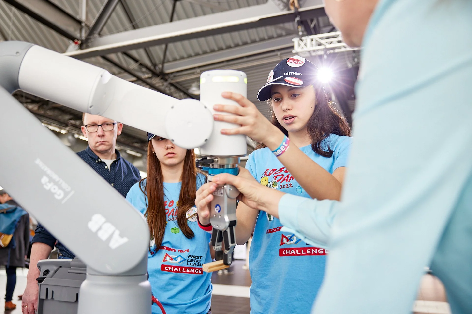 Junge Mädchen beim Experimentieren mit einem Roboterarm während eines FIRST LEGO League Events, unter Aufsicht eines Erwachsenen, in einer industriellen Umgebung.