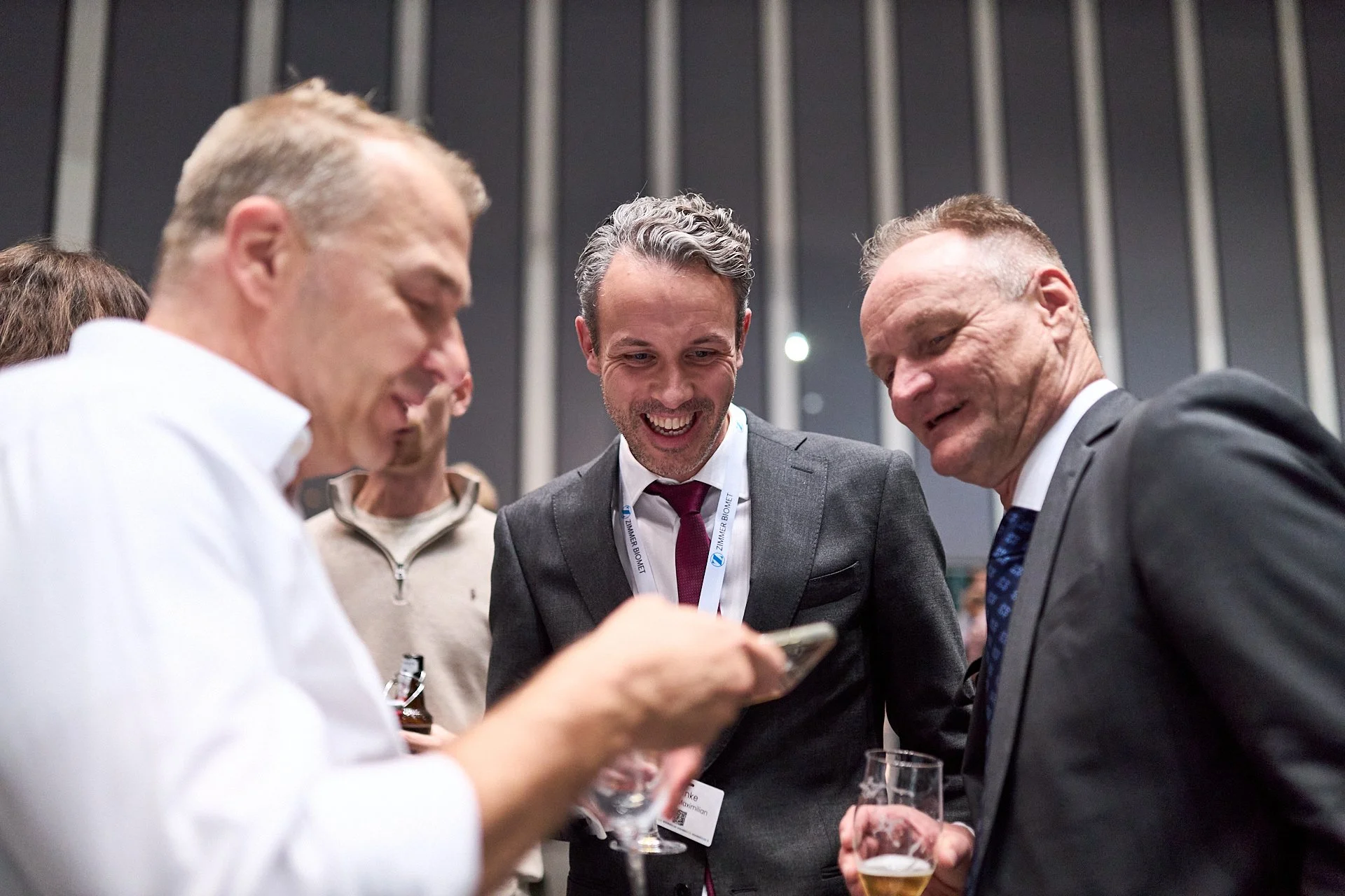 Vier Männer in formeller Kleidung lachen und schauen auf ein Smartphone, während sie bei einer Veranstaltung Getränke halten.