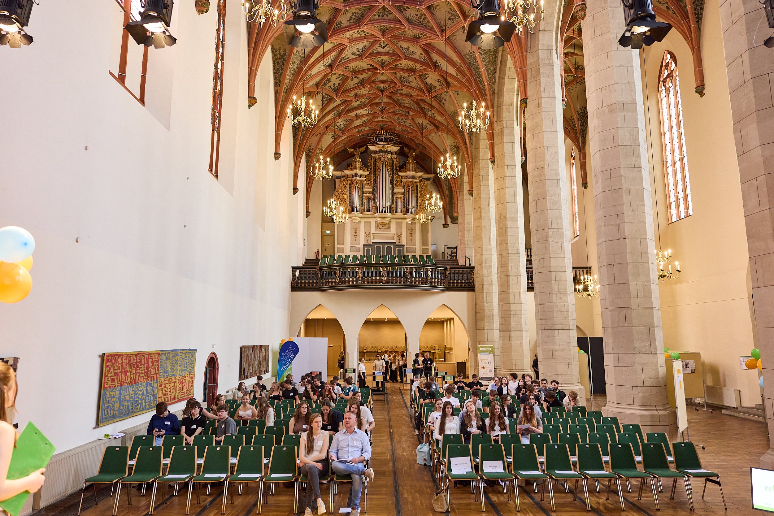 Ein großer Saal mit hohen, gewölbten Decken, in dem viele Menschen sitzen und warten, vermutlich bei einer Veranstaltung. Der Raum ist mit viktorianischer Architektur gestaltet, mit Säulen, großen Fenstern und einer Orgel im Hintergrund.