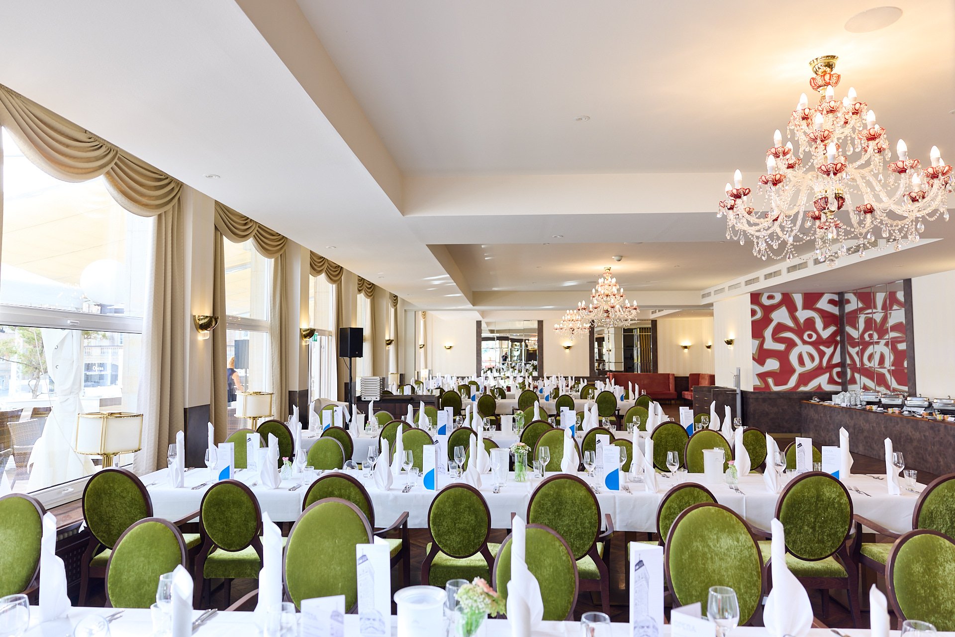 Ein festlich gedeckter Speisesaal mit weißen Tischen, grünen Stühlen, weißen Servietten, Felpgläsern und Menükarten. Der Raum hat große Fenster mit cremefarbenen Vorhängen und bietet elegante Kronleuchter sowie Wandlampen als Beleuchtung.