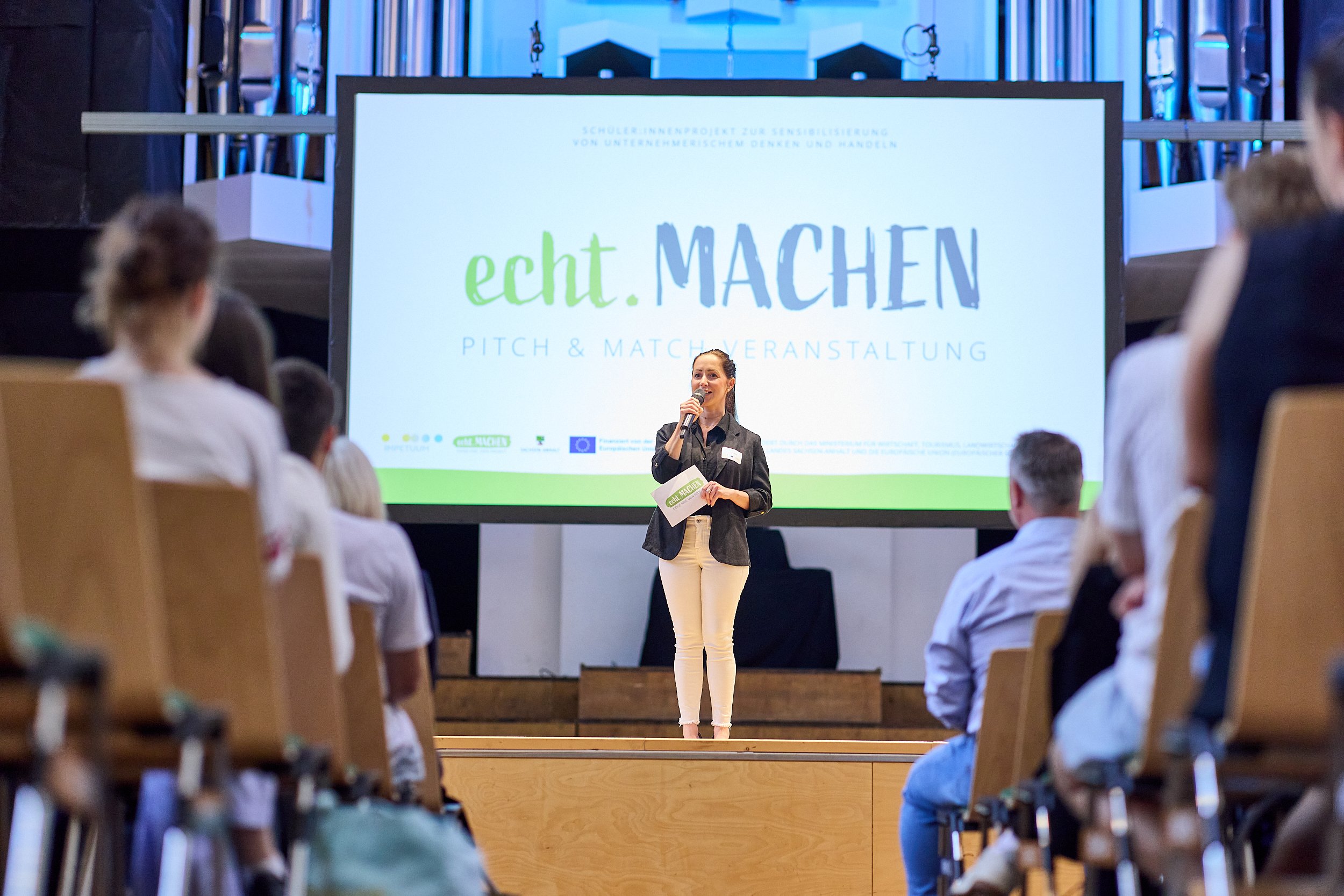 Eine Frau steht auf einer Bühne und spricht in ein Mikrofon vor einem Publikum bei einer Veranstaltung namens 'echte MACHE'.