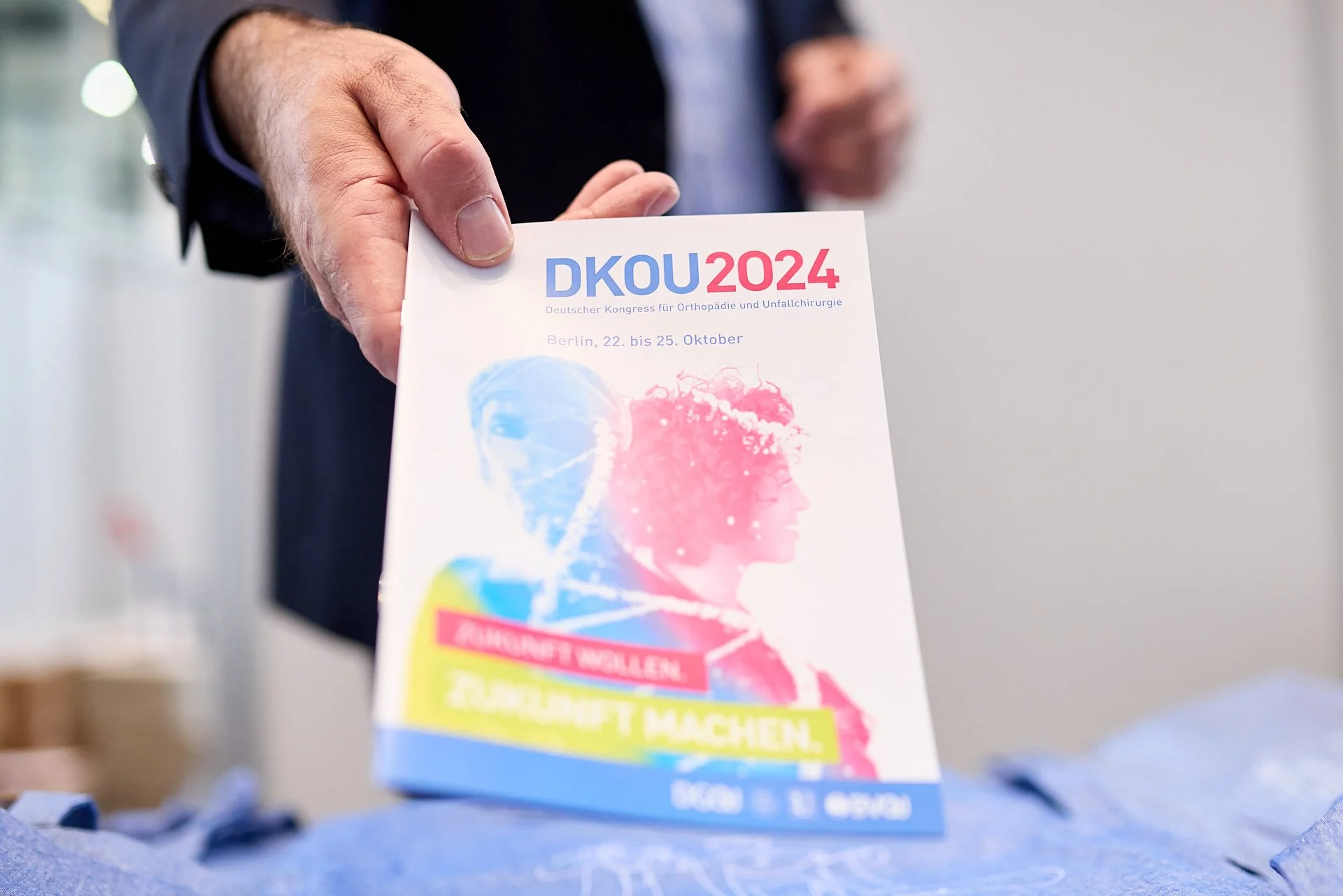 Hand hält eine Broschüre für den Deutschen Kongress für Orthopädie und Unfallchirurgie 2024 in Berlin, vom 22. bis 25. Oktober, mit buntem Design und Silhouetten von zwei Personen.