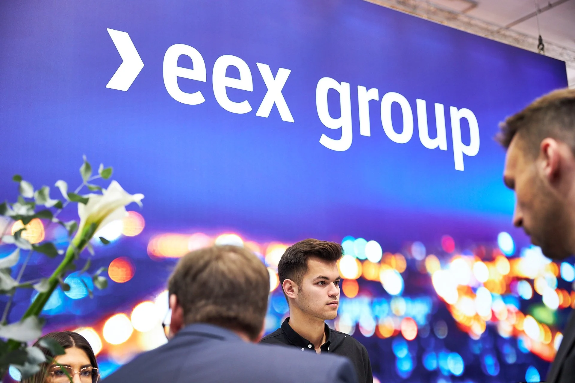 Menschen bei einer Veranstaltung vor einem großen Bildschirm mit dem Text ' eex group' auf einem blauen Hintergrund.