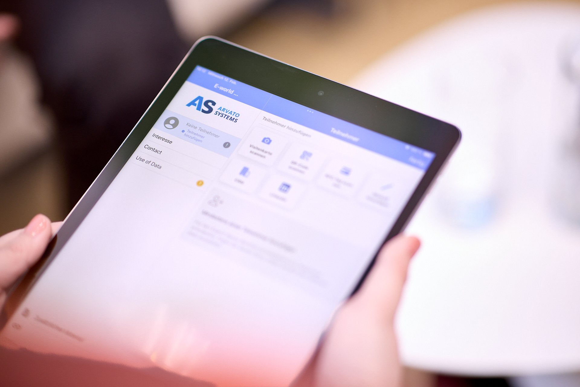 Eine Hand hält ein Tablet, das eine digitale Plattform namens 'AS ARAVATO SYSTEMS' zeigt, mit Menüoptionen und Symbolen für verschiedene Funktionen.