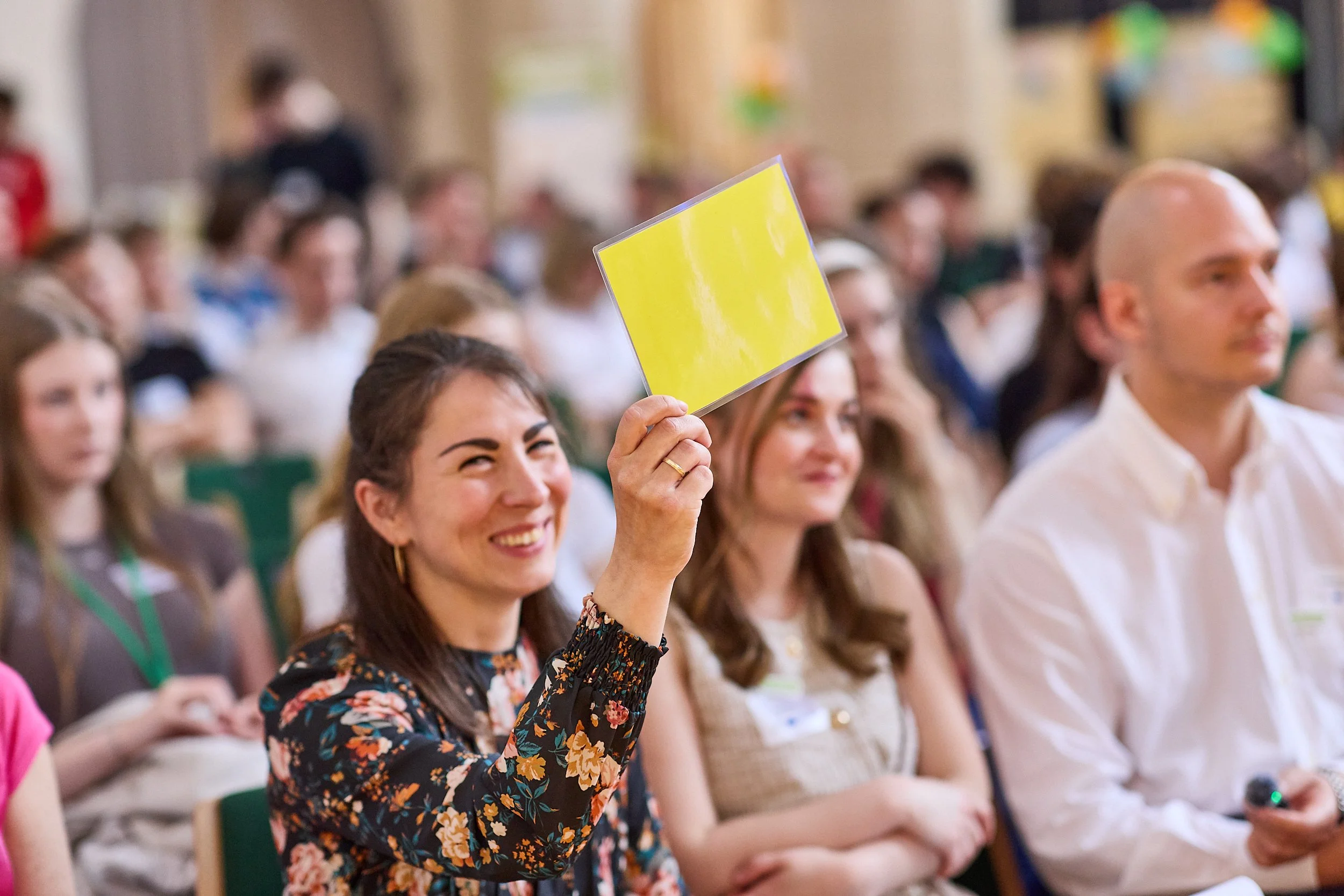 Eine Frau in einem Blumenmuster-Kleid hält ein gelbes Schild hoch, während sie in einem Publikum bei einer Veranstaltung sitzt.