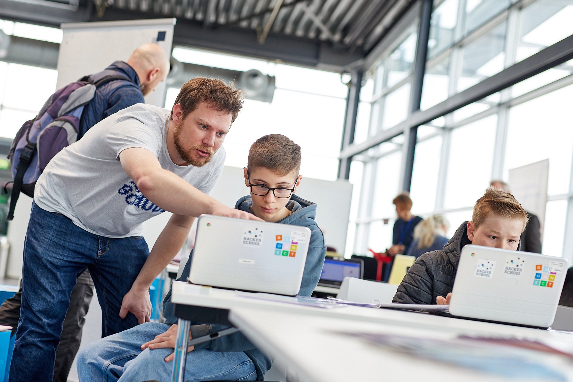 Ein Erwachsener zeigt einem Jungen an einem Laptop im Klassenzimmer, während ein weiterer Junge an einem anderen Laptop arbeitet.