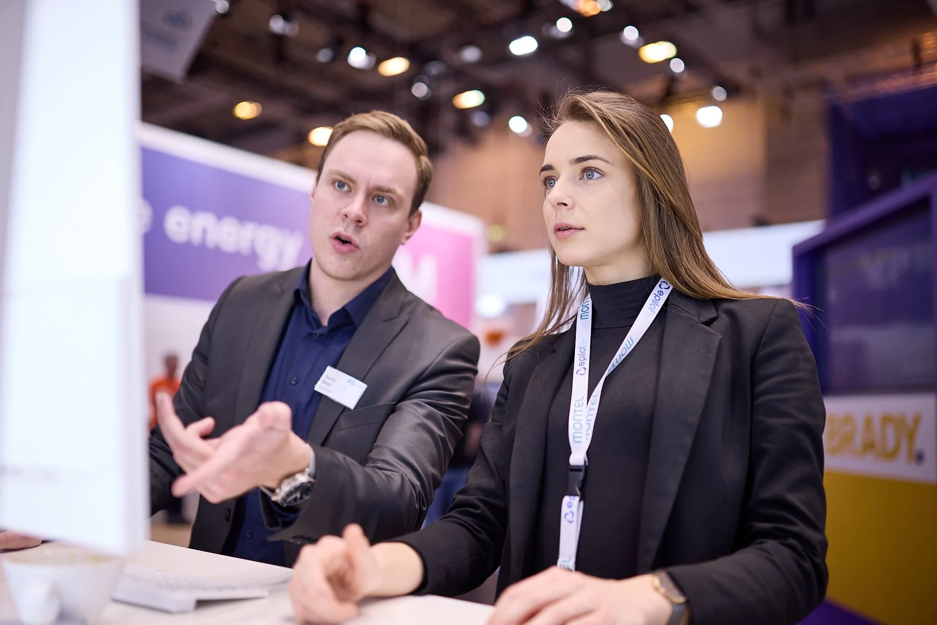 Zwei berufstätige Menschen im Gespräch bei einer Messe oder Konferenz, im Hintergrund Banner und leuchtende Deckenbeleuchtung.