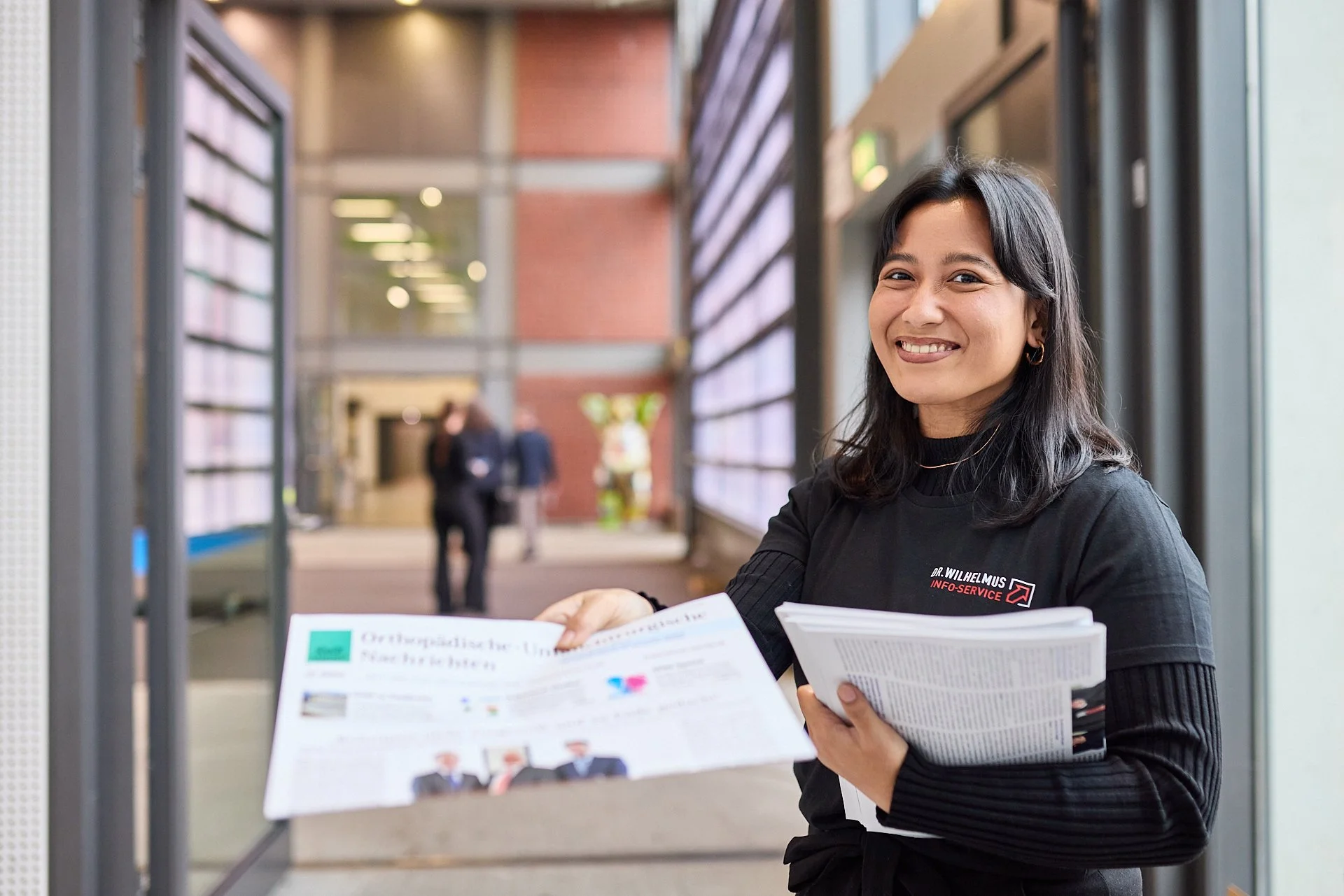 Eine Frau in schwarzer Kleidung hält Broschüren in einer modernen, hell erleuchteten Eingangshalle und lächelt in die Kamera.