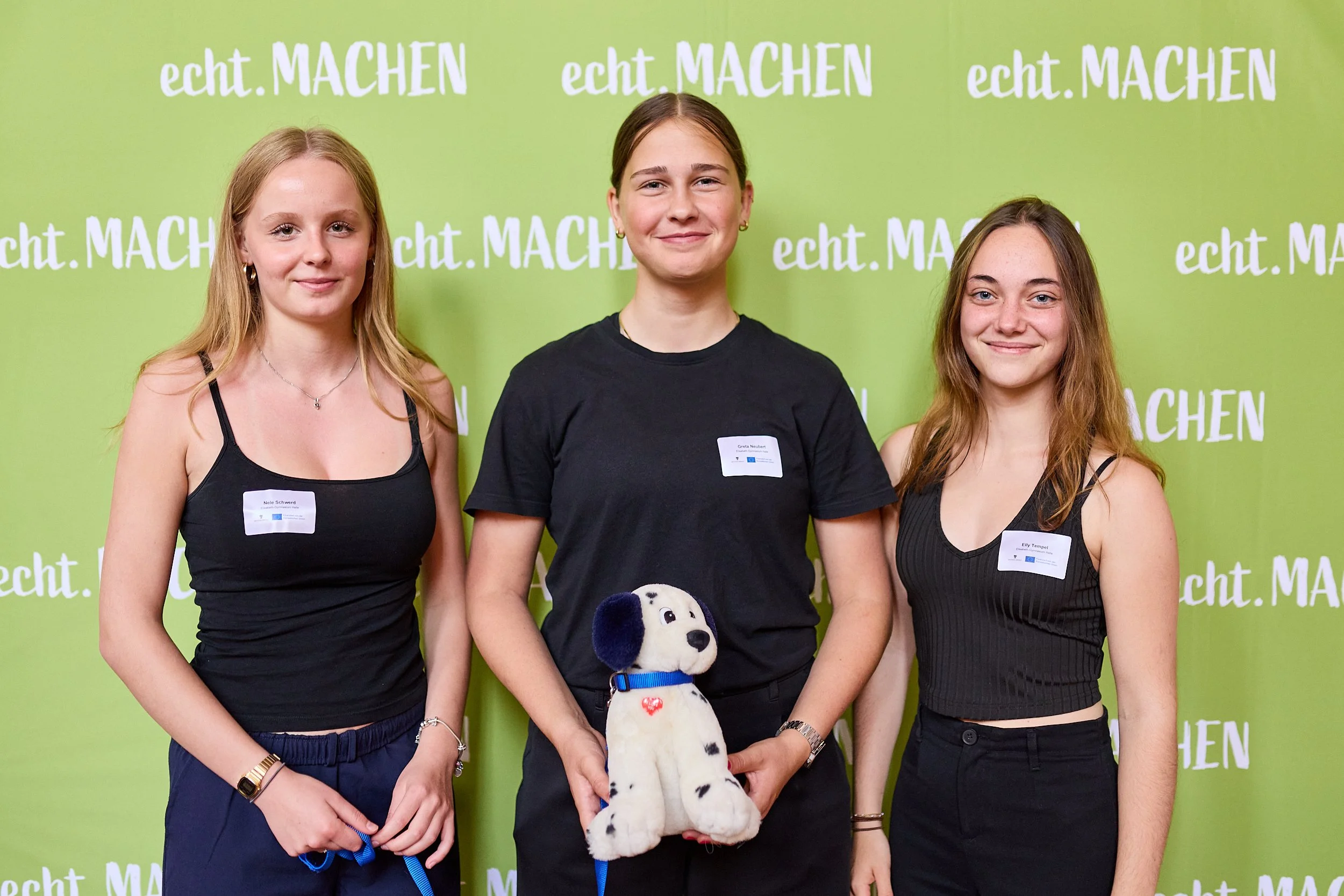 Drei junge Frauen stehen vor einem grünen Hintergrund mit dem Text "echt.MACHEN". Die Frau in der Mitte hält ein Plüschtier, einen Dalmatiner-Hund.