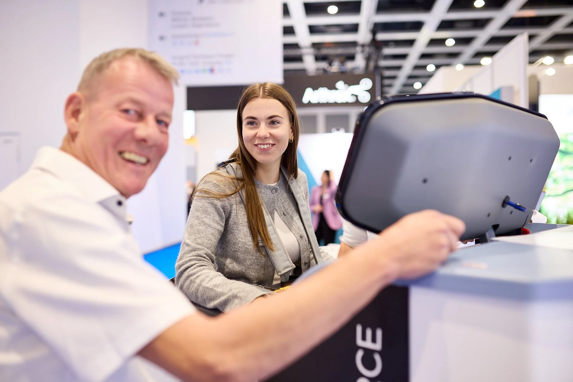 Zwei Menschen an einem Messestand, lächelnd, ein Mann in Weiß, eine junge Frau in grauem Pullover, im Hintergrund eine Messehalle.