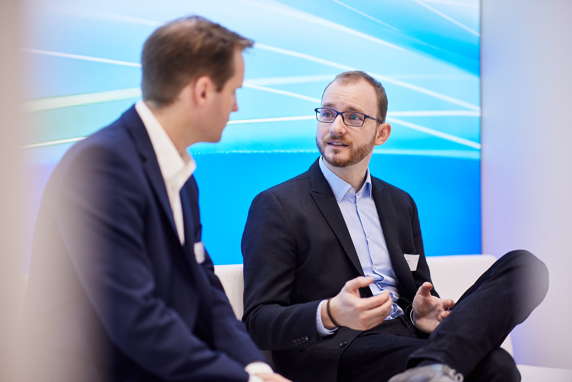 Zwei Männer im Gespräch, einer mit Brille und dunklem Anzug, der andere mit dunkler Jacke, in einem modernen Raum mit blauer Wand im Hintergrund.